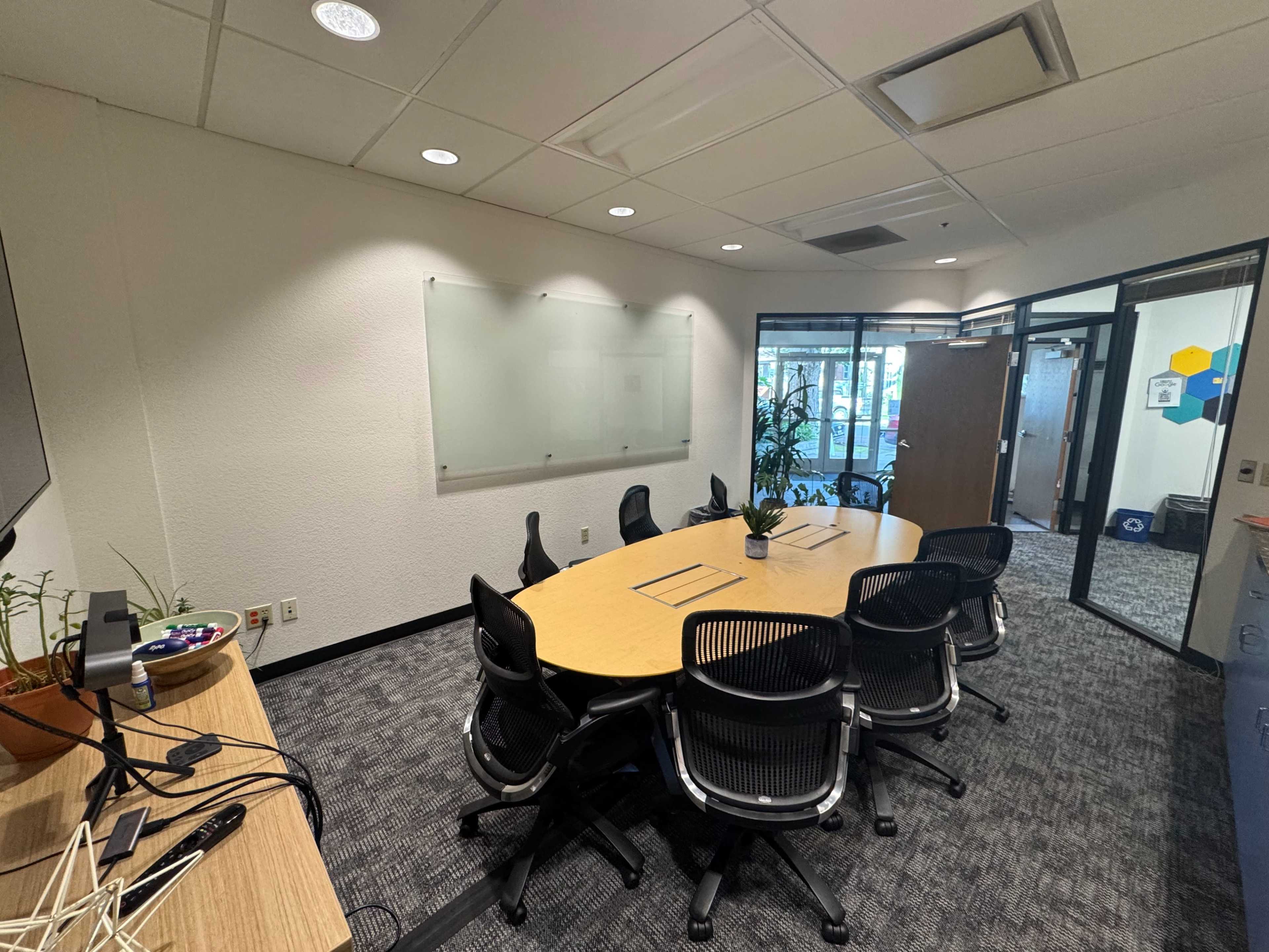 The image shows a meeting room with a large round table surrounded by black chairs, a whiteboard on one wall, and glass doors leading to another area.