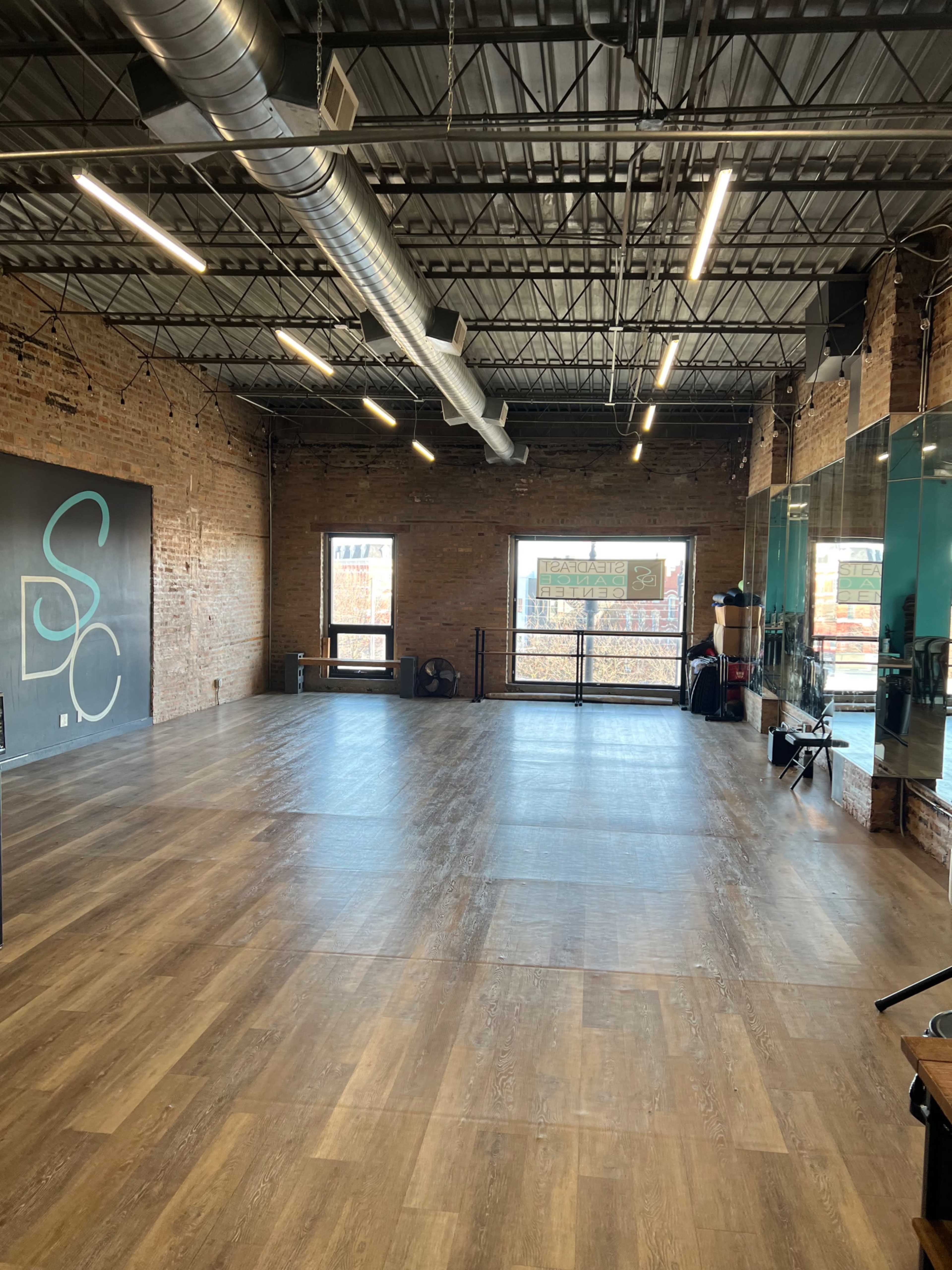A spacious studio with wooden flooring, exposed brick walls, large windows, and mirrors on one side.