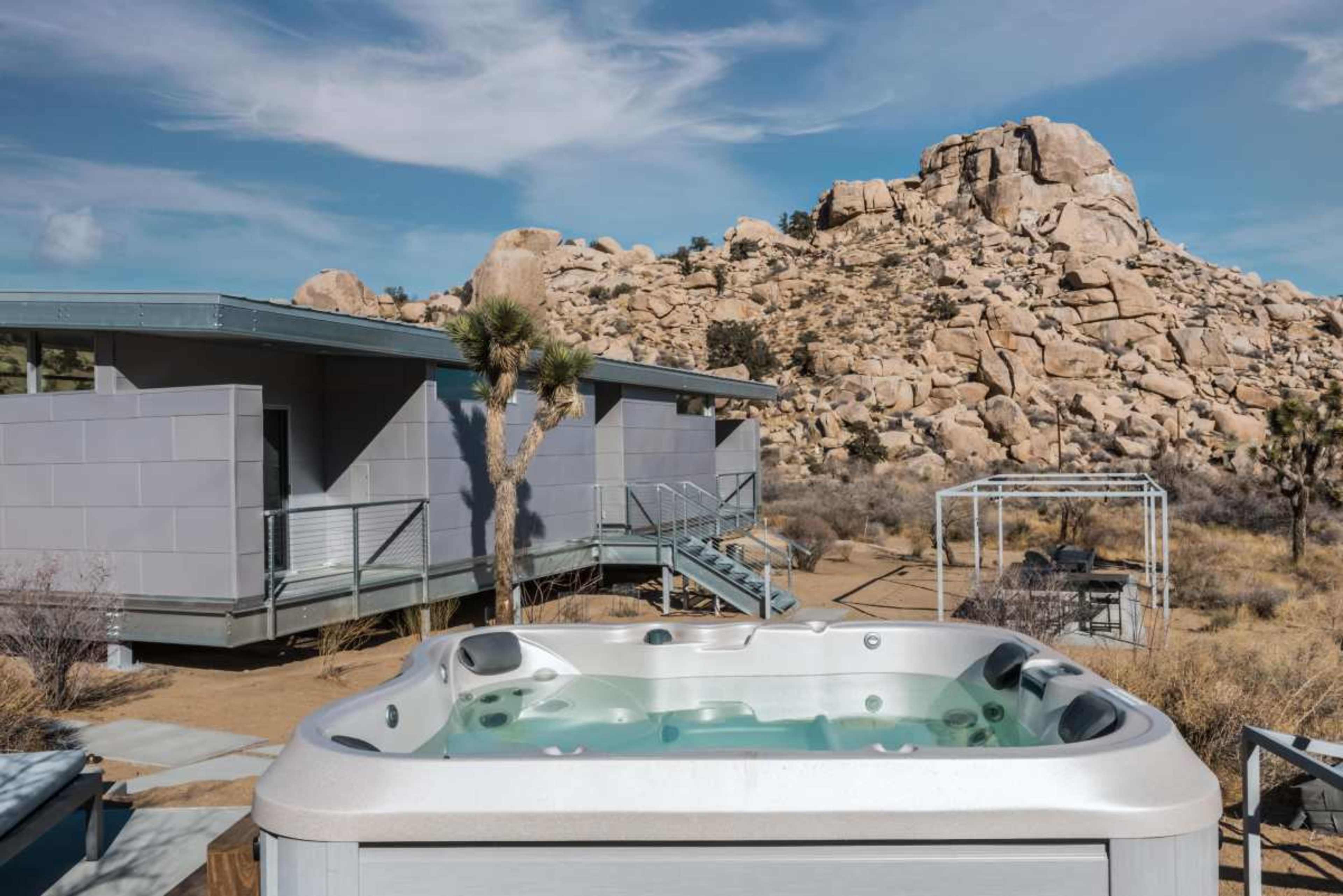 A hot tub is situated in the foreground with a rocky hillside and a modern building in the background.