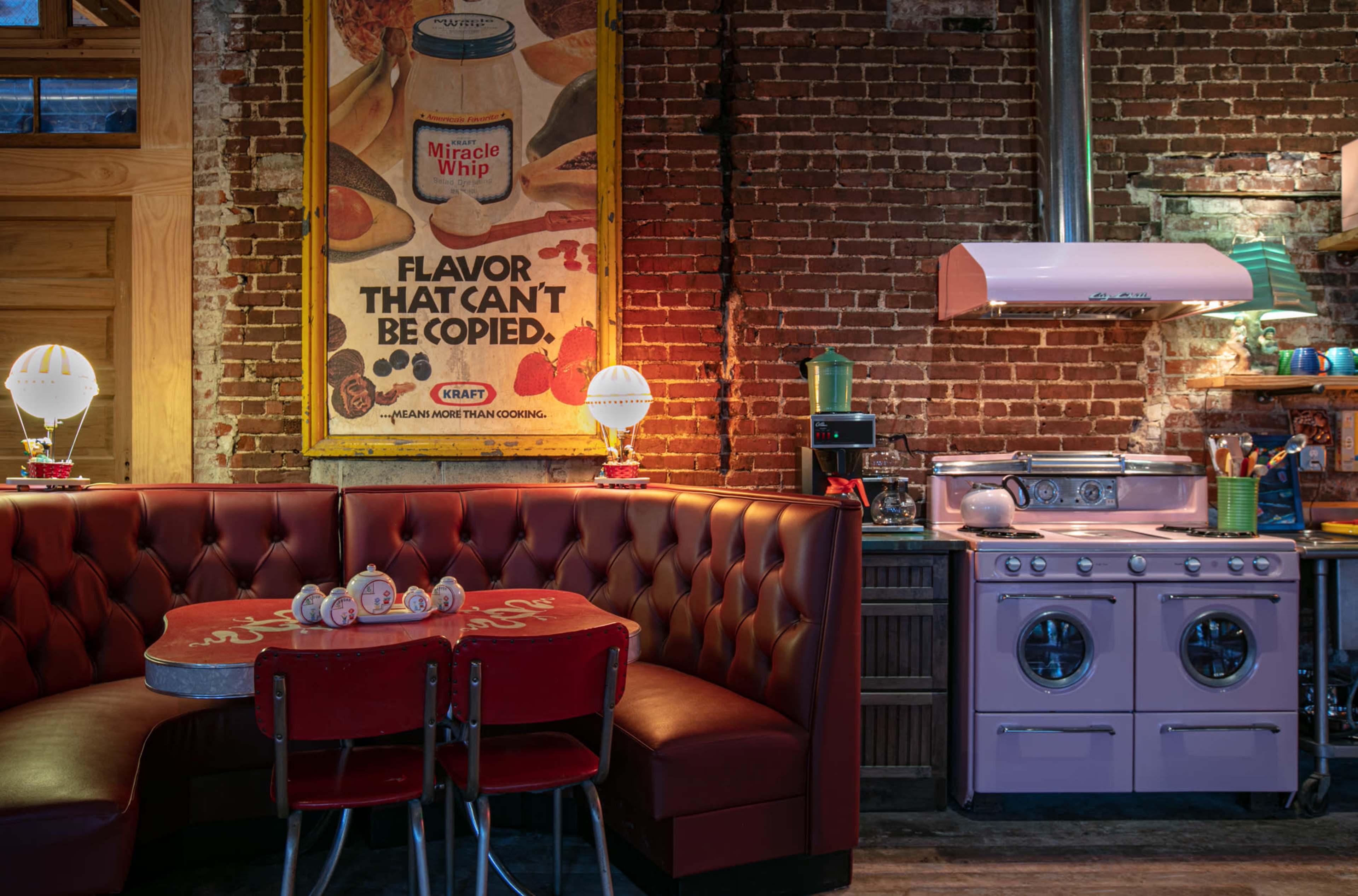 A retro kitchen with a pink stove and a cozy booth area featuring a vintage Miracle Whip advertisement on the brick wall.