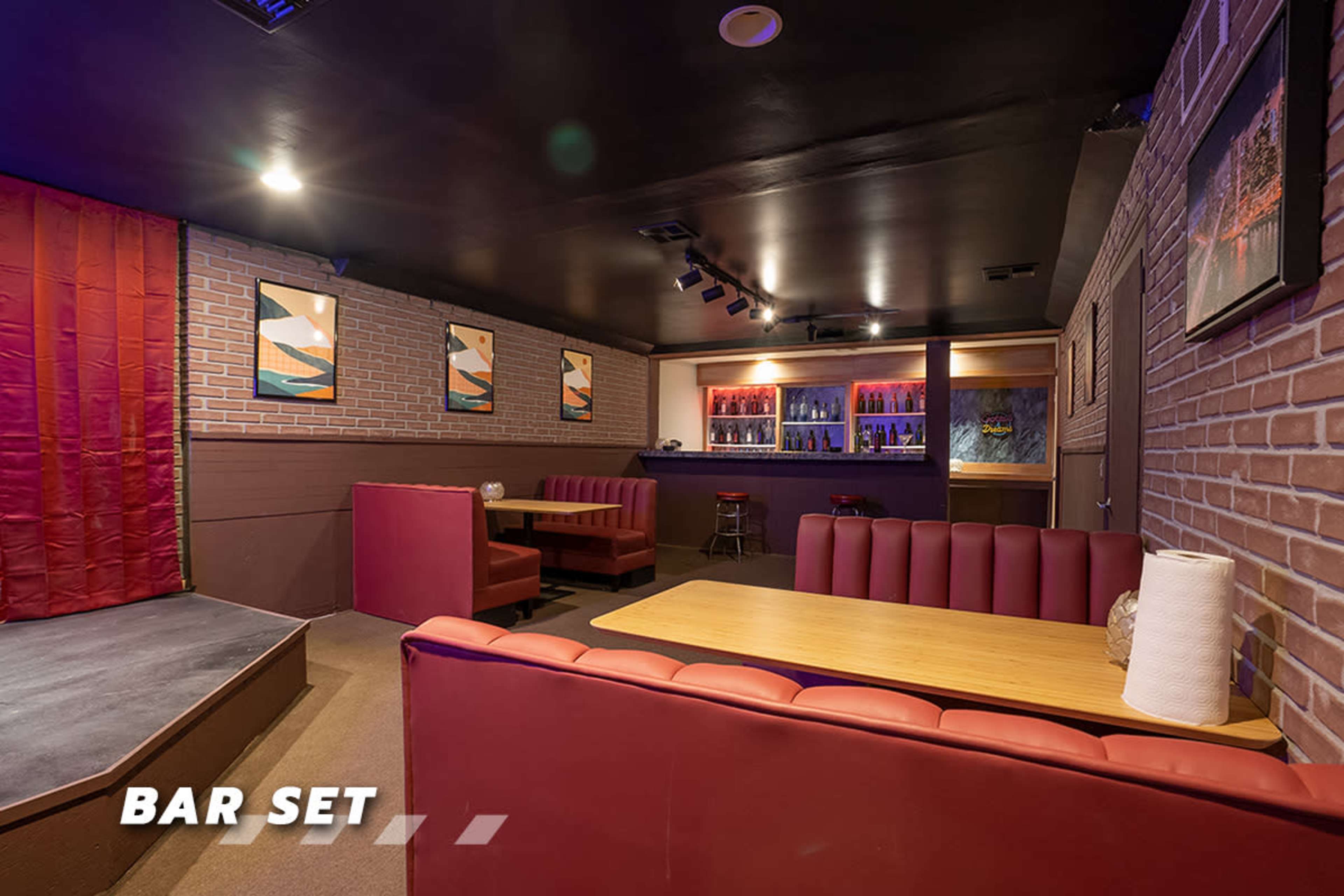 The image shows a bar set with red seating, wooden tables, and a backlit bar area featuring various bottles, set against a brick wall.