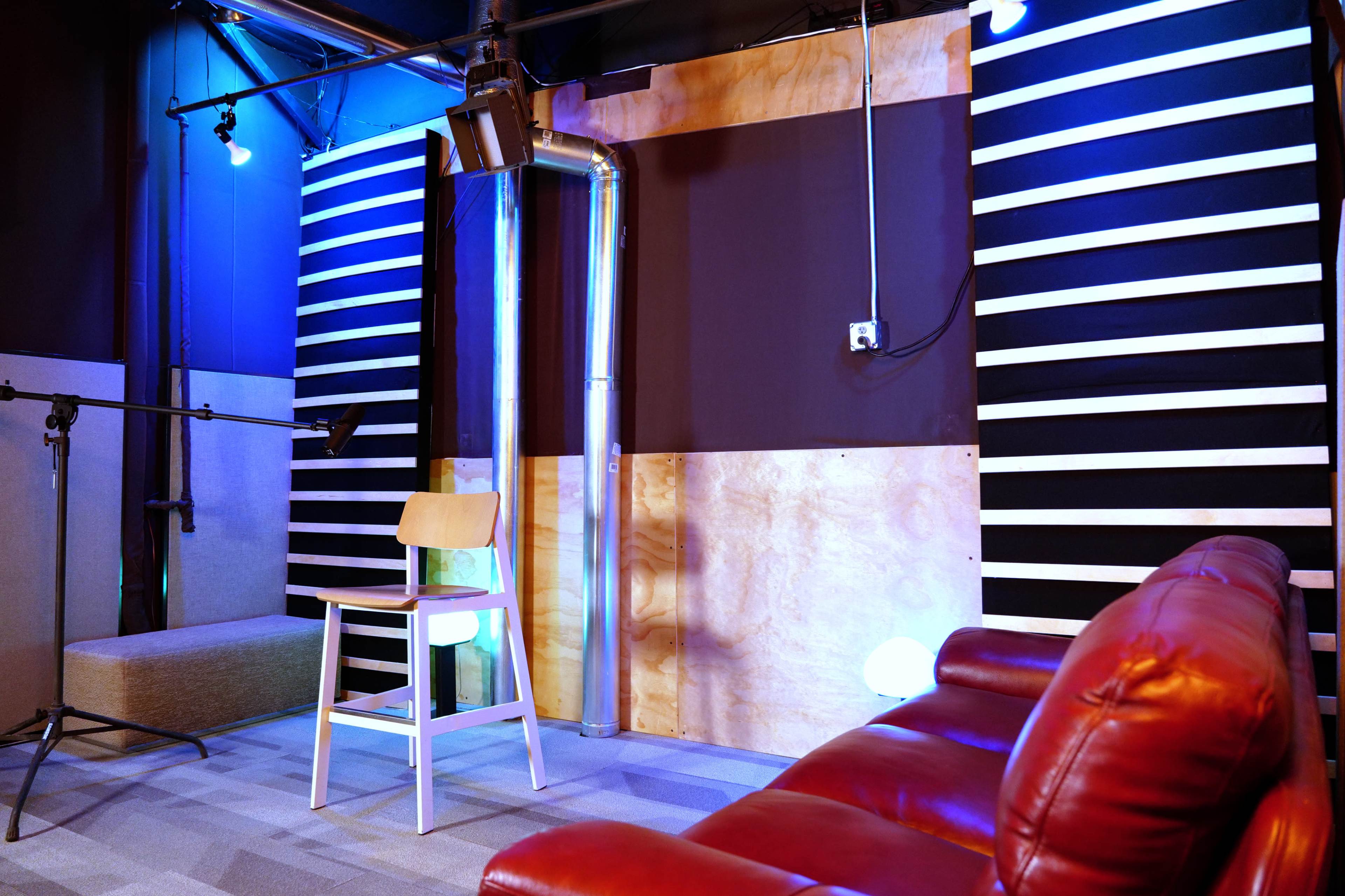 A small recording studio with a red sofa, a wooden chair, and a microphone stand against a backdrop of black and wooden panels.