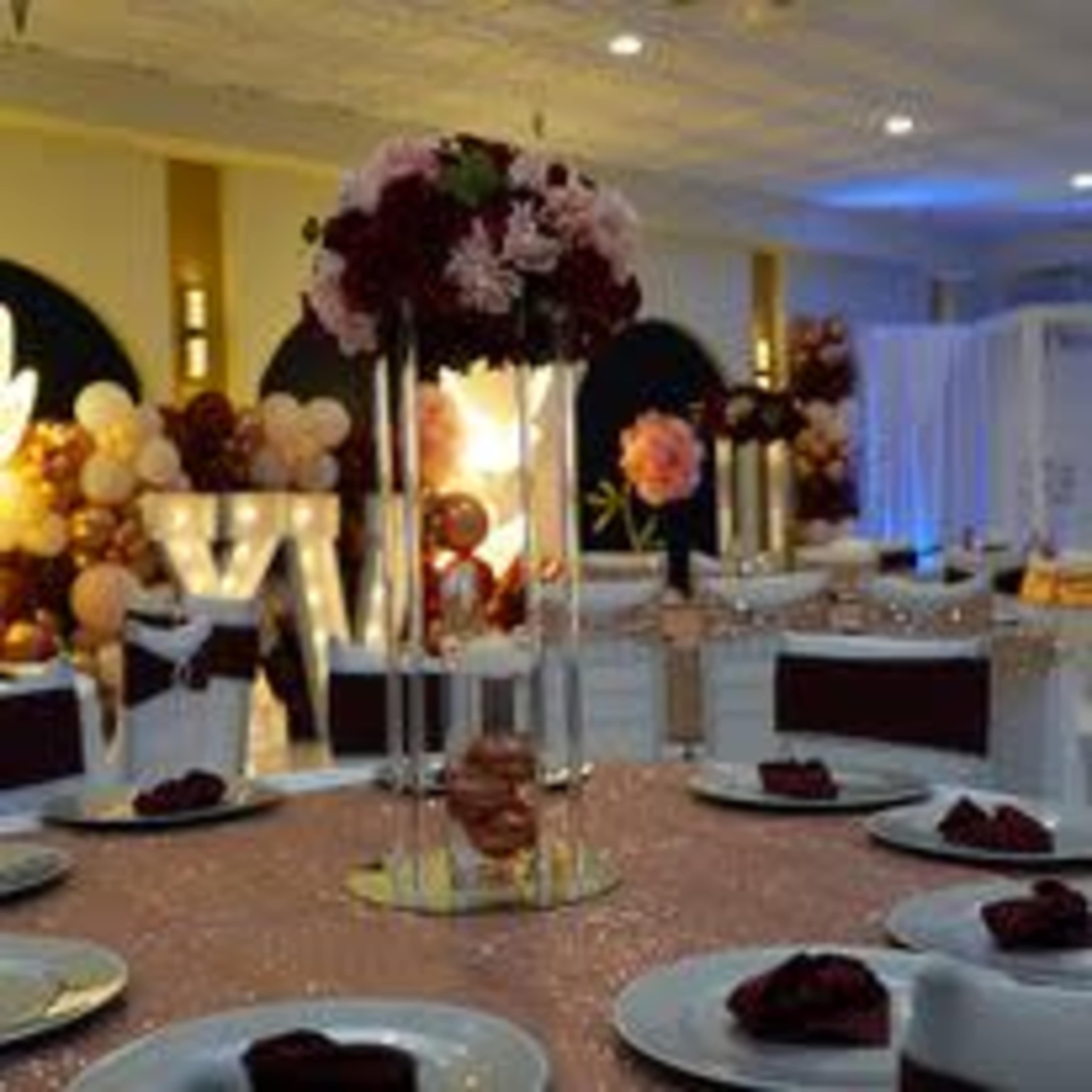 A decorated banquet hall with tables set for an event, featuring floral centerpieces and balloons in the background.