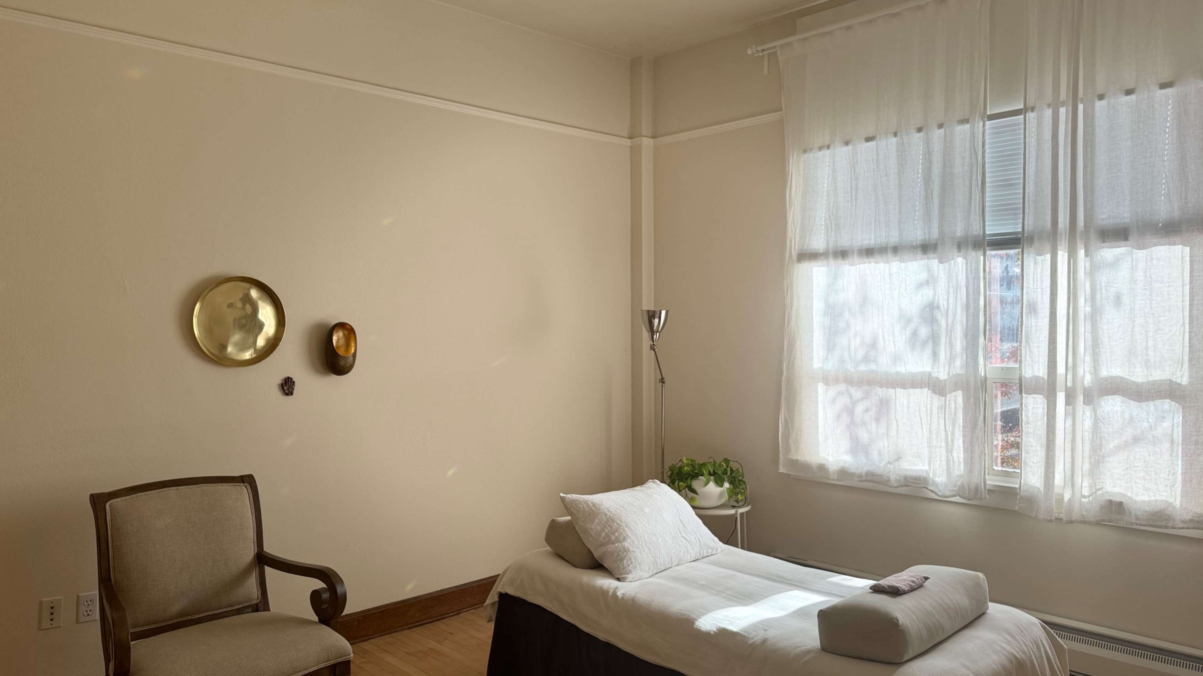 The image shows a serene, minimalist room with a massage table, a chair, and a window draped with sheer curtains allowing light to filter in.