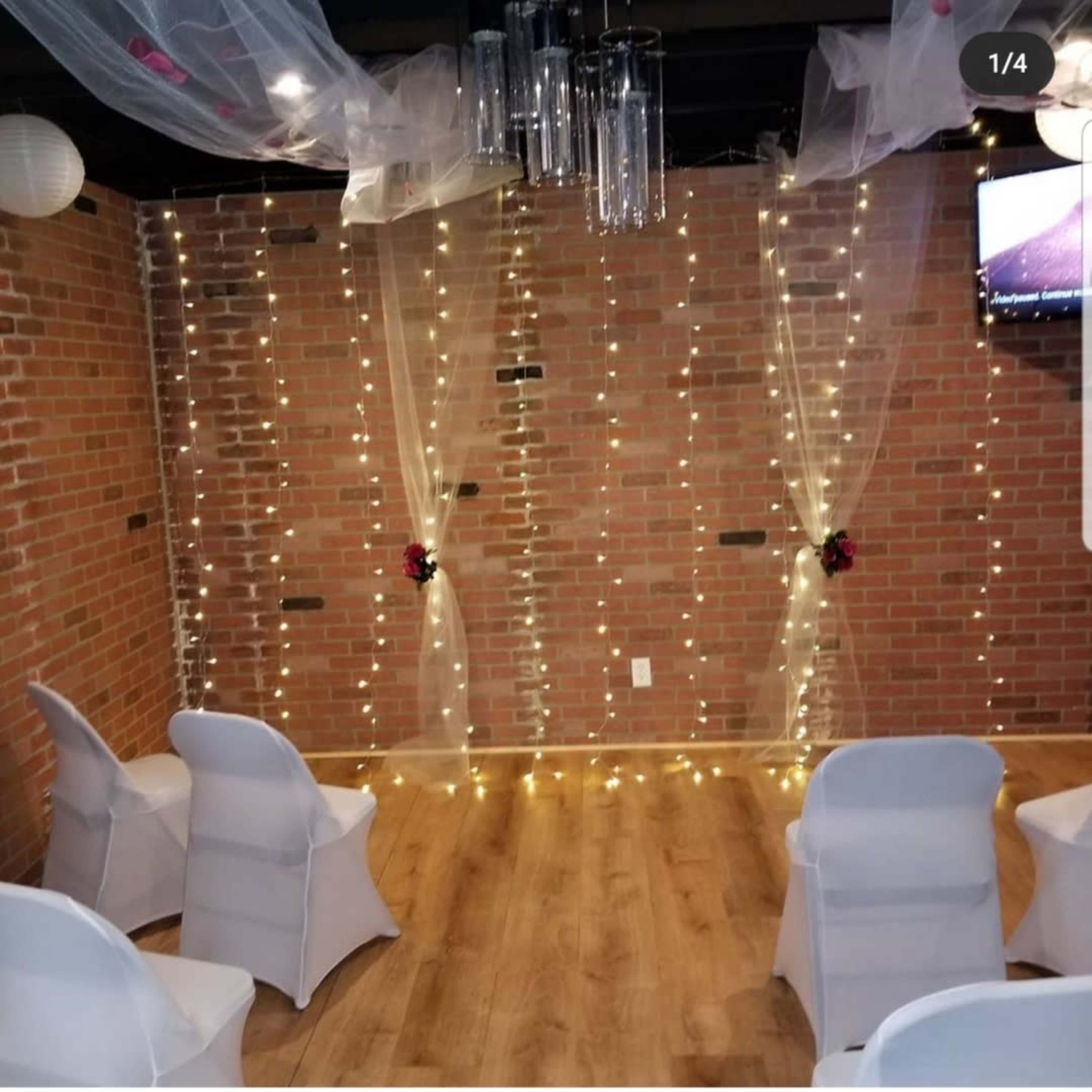 The scene features a decorated interior space with white chairs arranged in front of a brick wall adorned with string lights and sheer drapes.