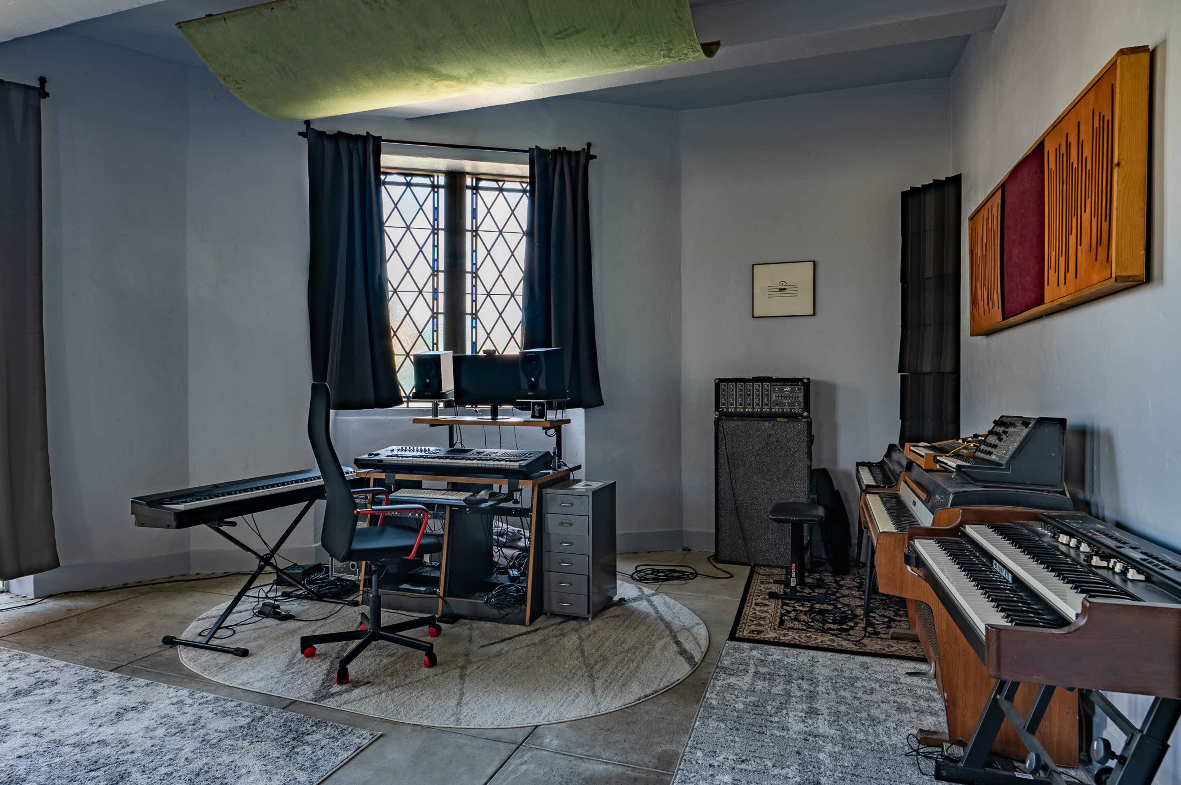 A music production room features a keyboard, computer setup, amplifier, and various musical instruments against a backdrop of windows and curtains.