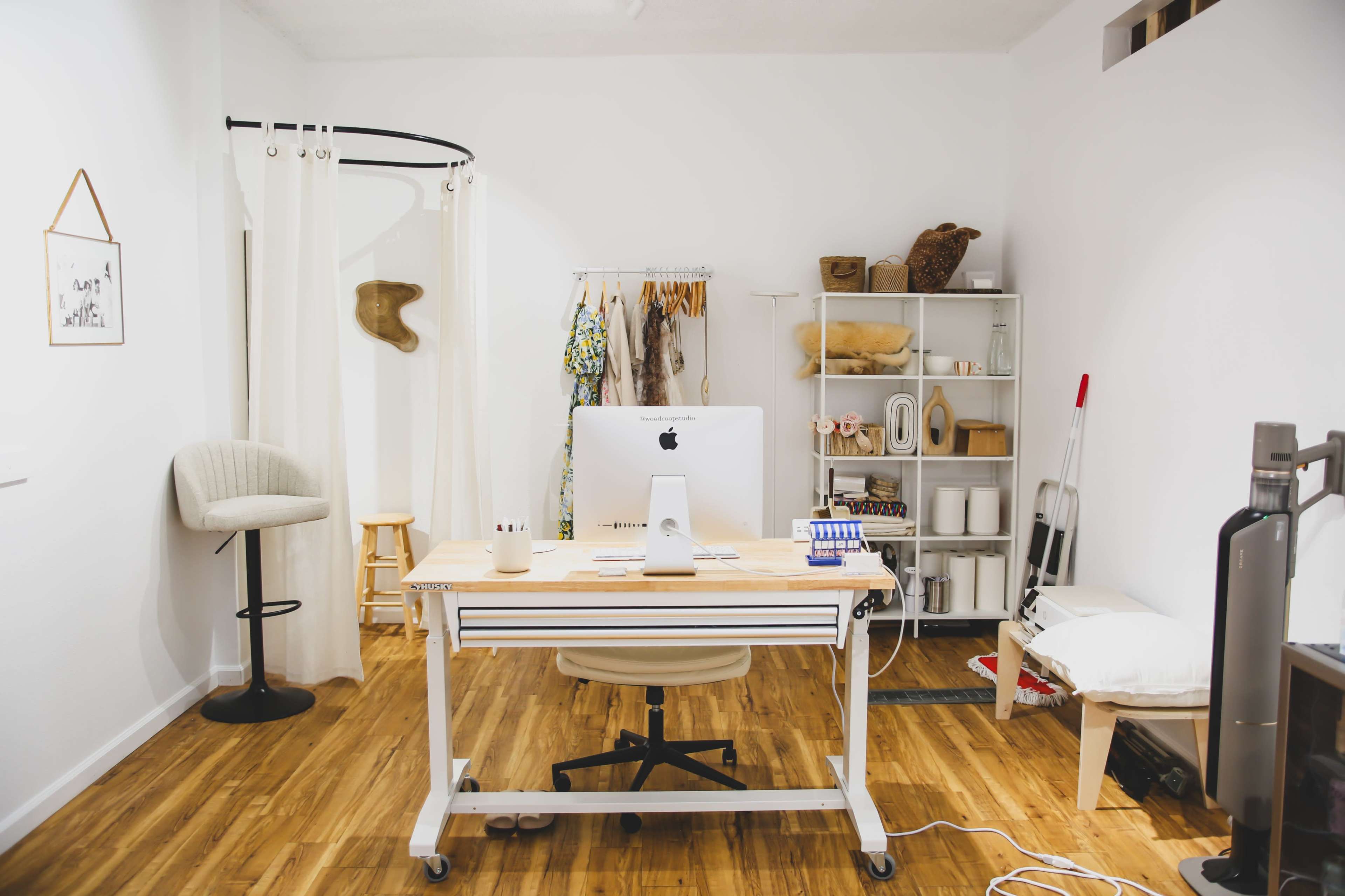 The image shows a minimalist office space featuring a desk with an iMac, a stool, clothing racks, and various organizational shelves against a plain white wall.