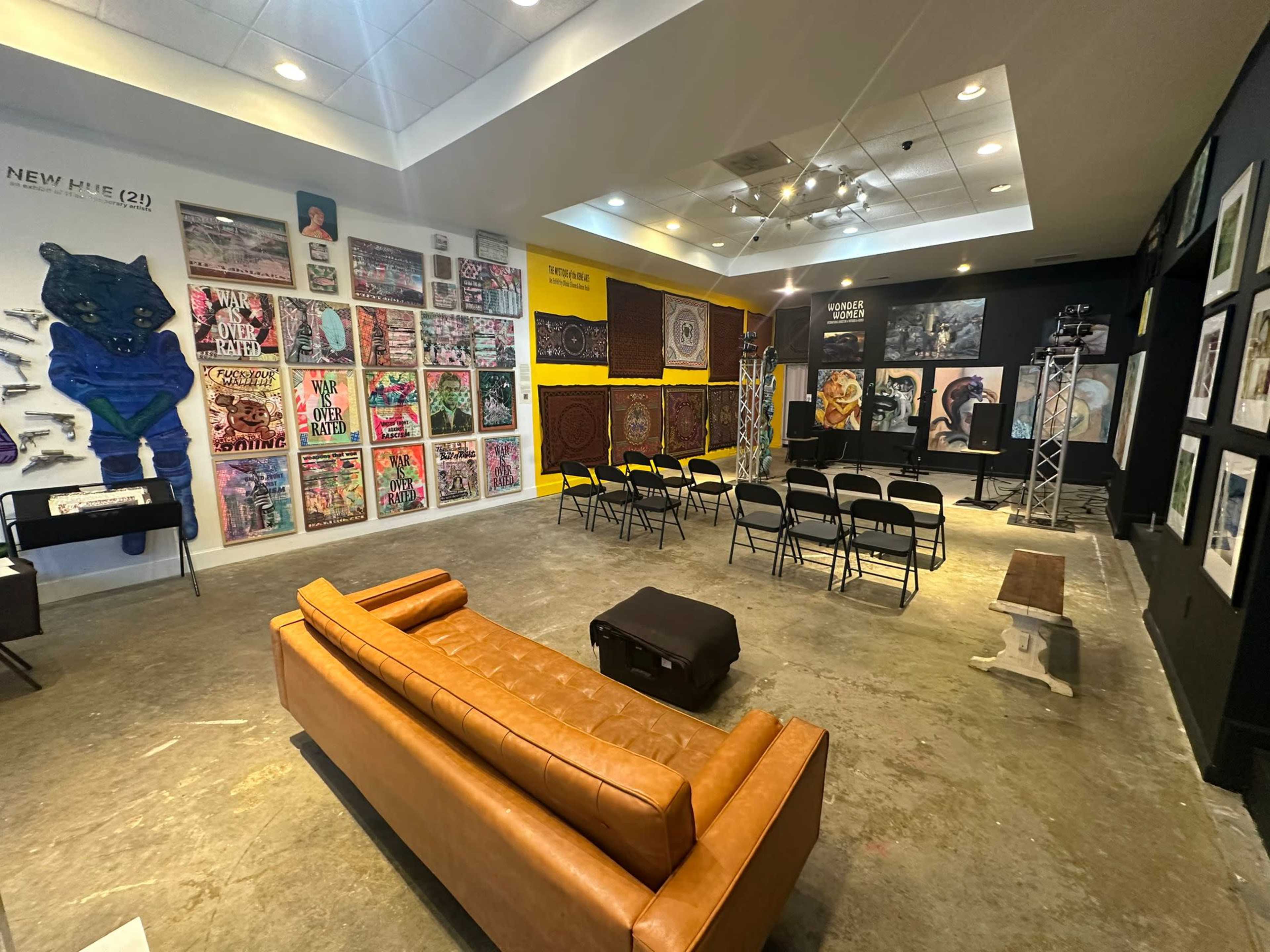 An art gallery or exhibition space featuring various wall-mounted artworks, a seating arrangement with black chairs, and a brown sofa in the foreground.