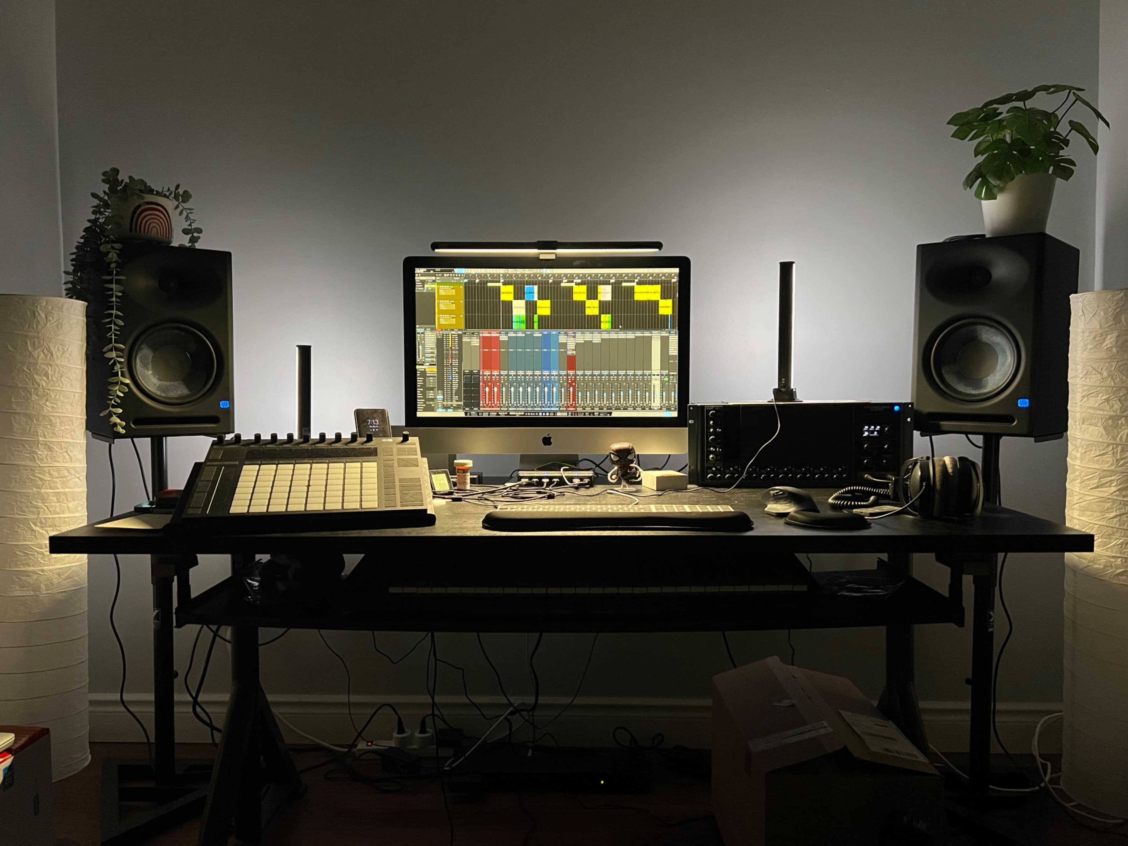 A music production setup featuring a computer monitor, audio speakers, a MIDI controller, headphones, and ambient lighting on a desk.