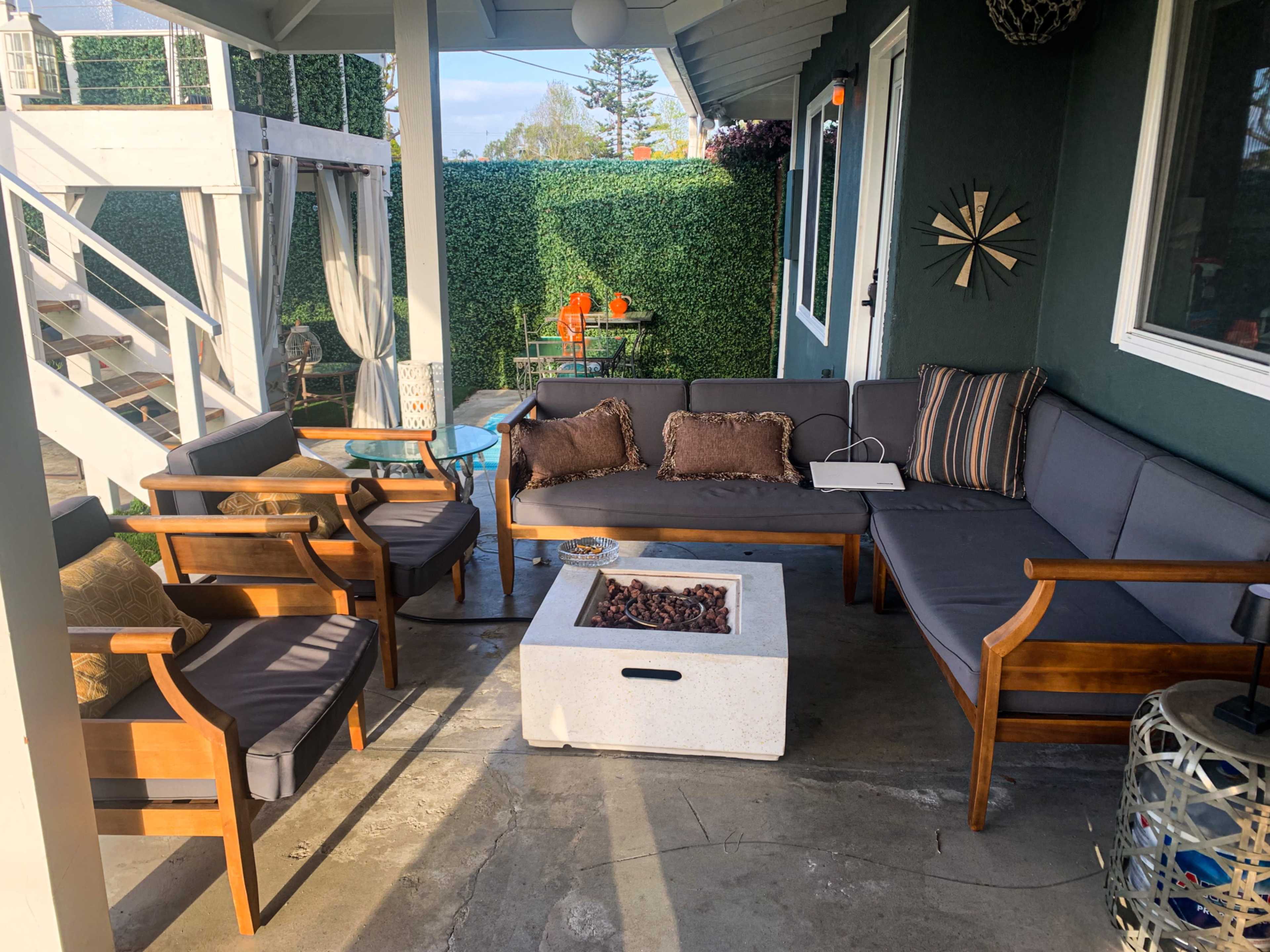 The image shows an outdoor seating area featuring a sectional sofa, wooden chairs, and a modern fire pit on a concrete floor, surrounded by greenery.