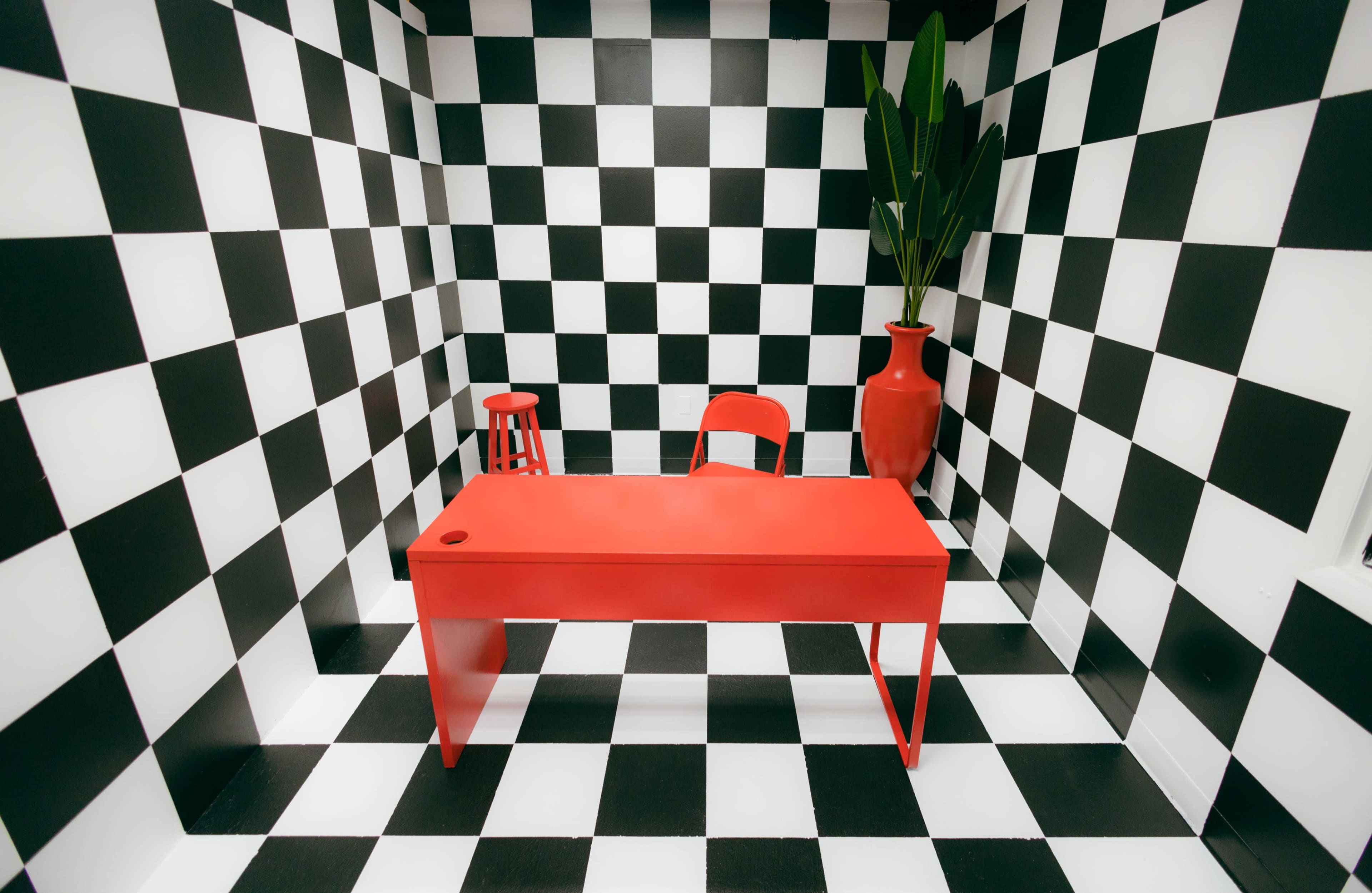 A red table and chair are positioned in a room with black and white checkered walls and floor, accompanied by a potted plant and a red stool.