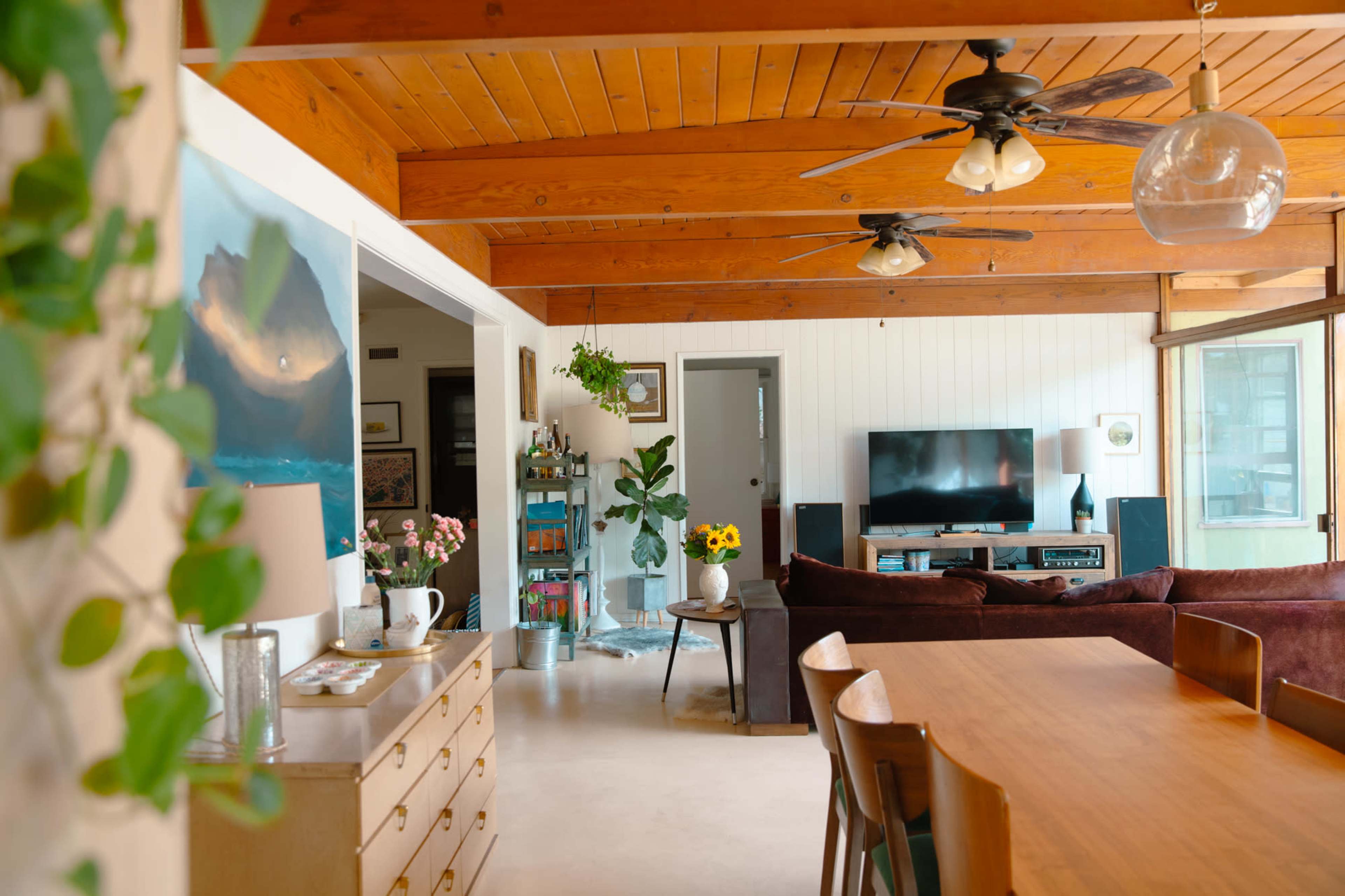 A spacious living area features wooden beams, a dining table, a television, and plants, with a view into a bright kitchen area.