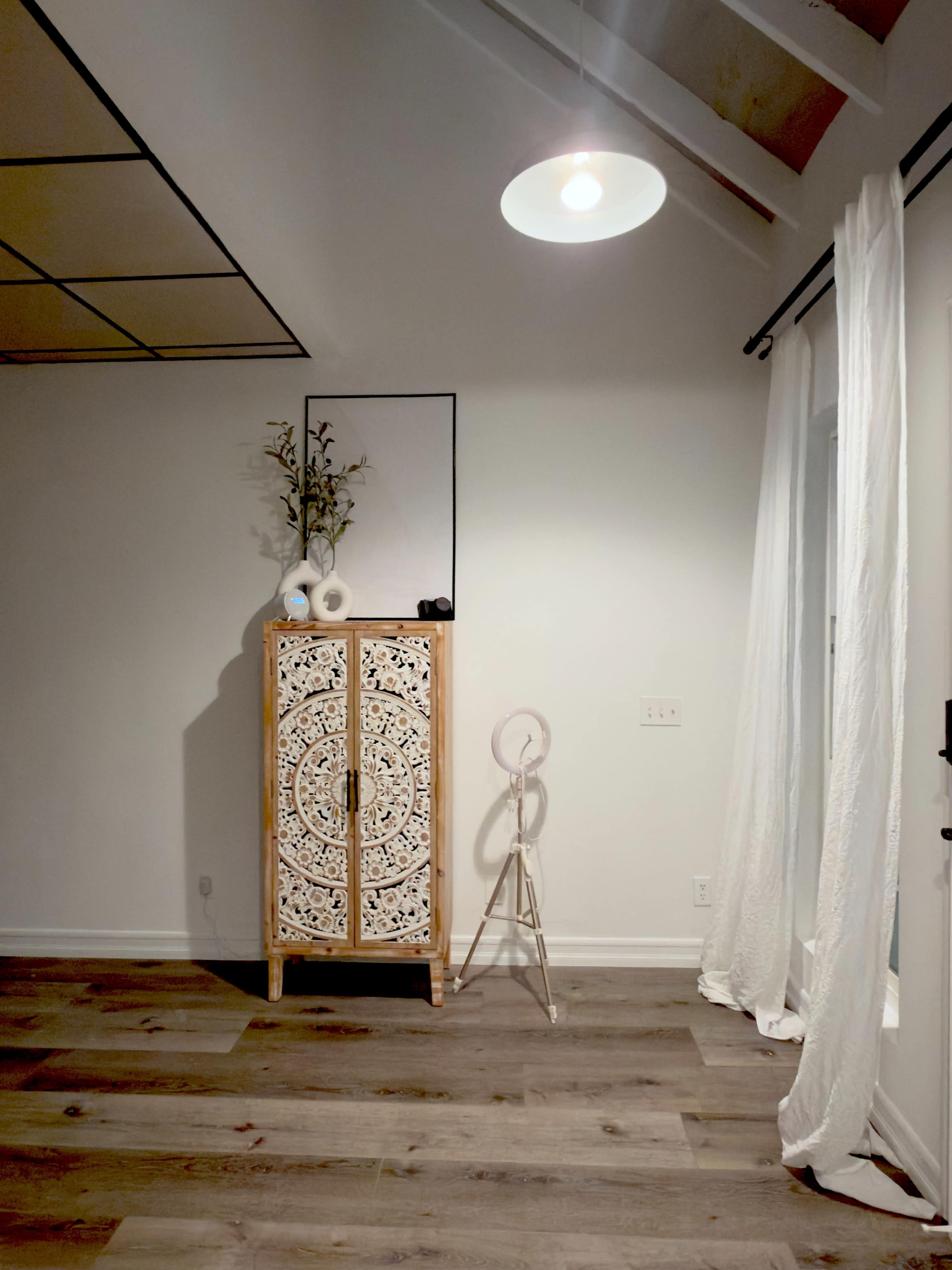 The image shows a softly lit room featuring a decorative wooden cabinet with intricate patterns, a floor lamp, and sheer white curtains.