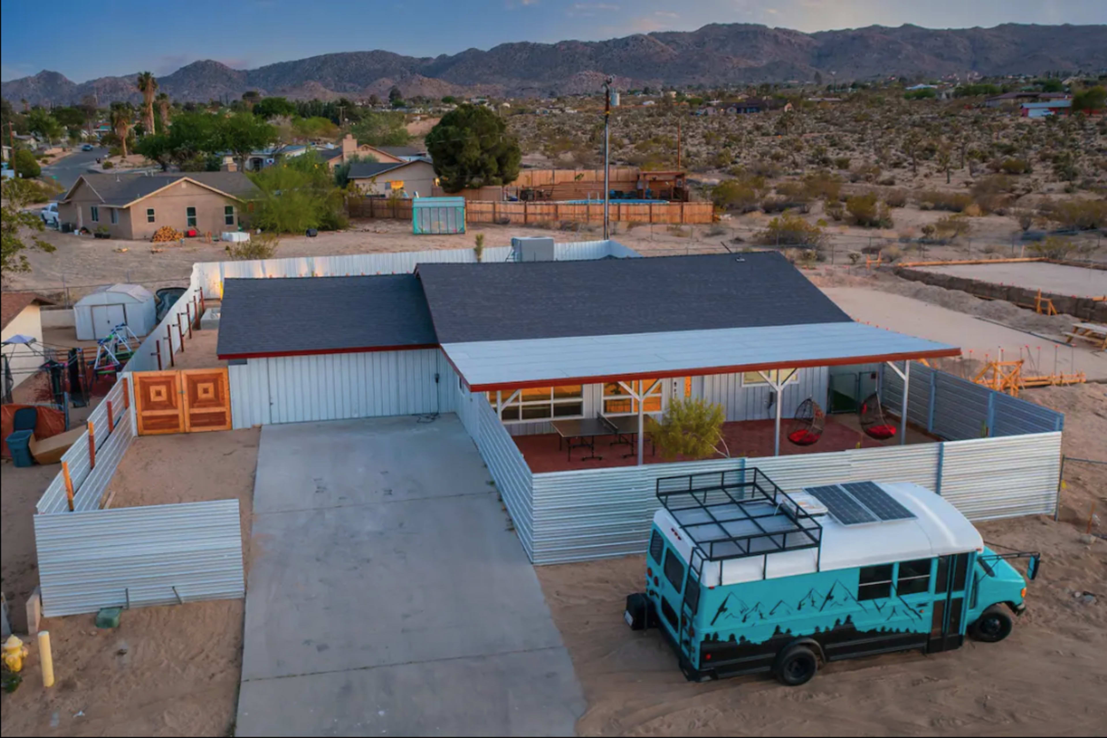 A modern metal-clad house with a fenced patio is situated in a desert landscape, surrounded by mountains and sparse vegetation.