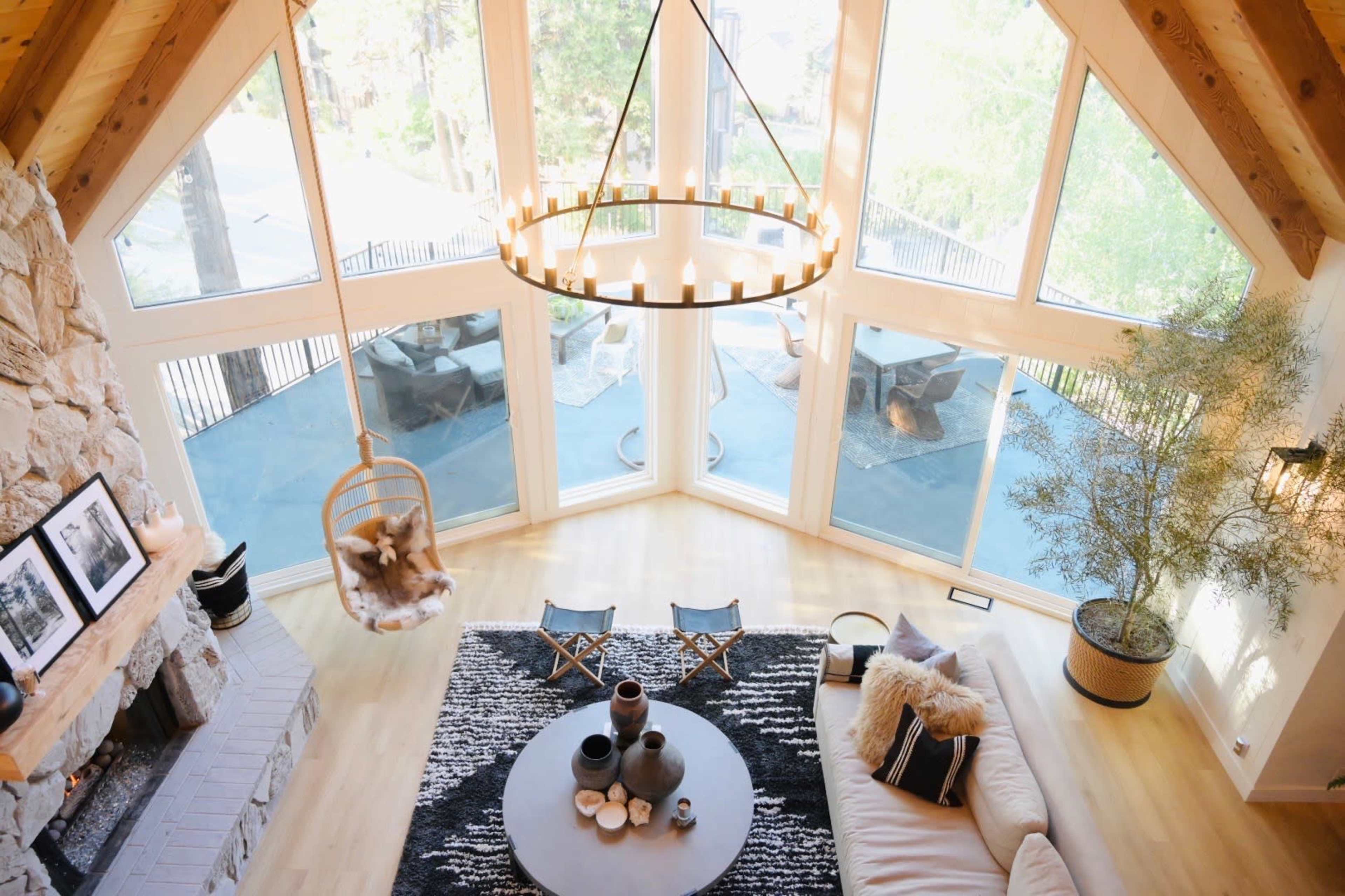 A spacious living room with large windows, a stone fireplace, a circular chandelier, and modern furnishings, including a hanging chair and a central coffee table.