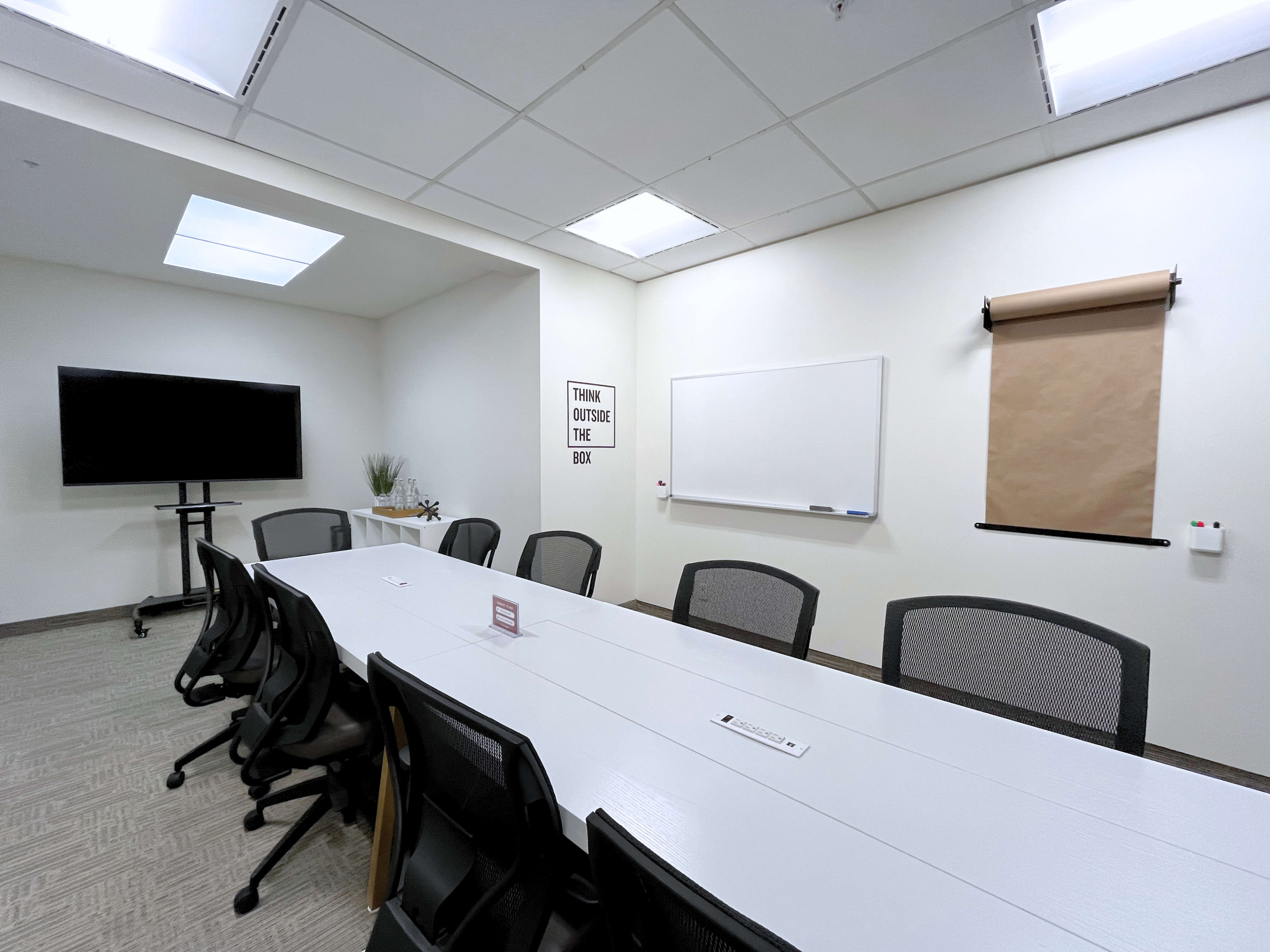 A modern conference room features a long white table surrounded by black chairs, a whiteboard, and a television on one wall.
