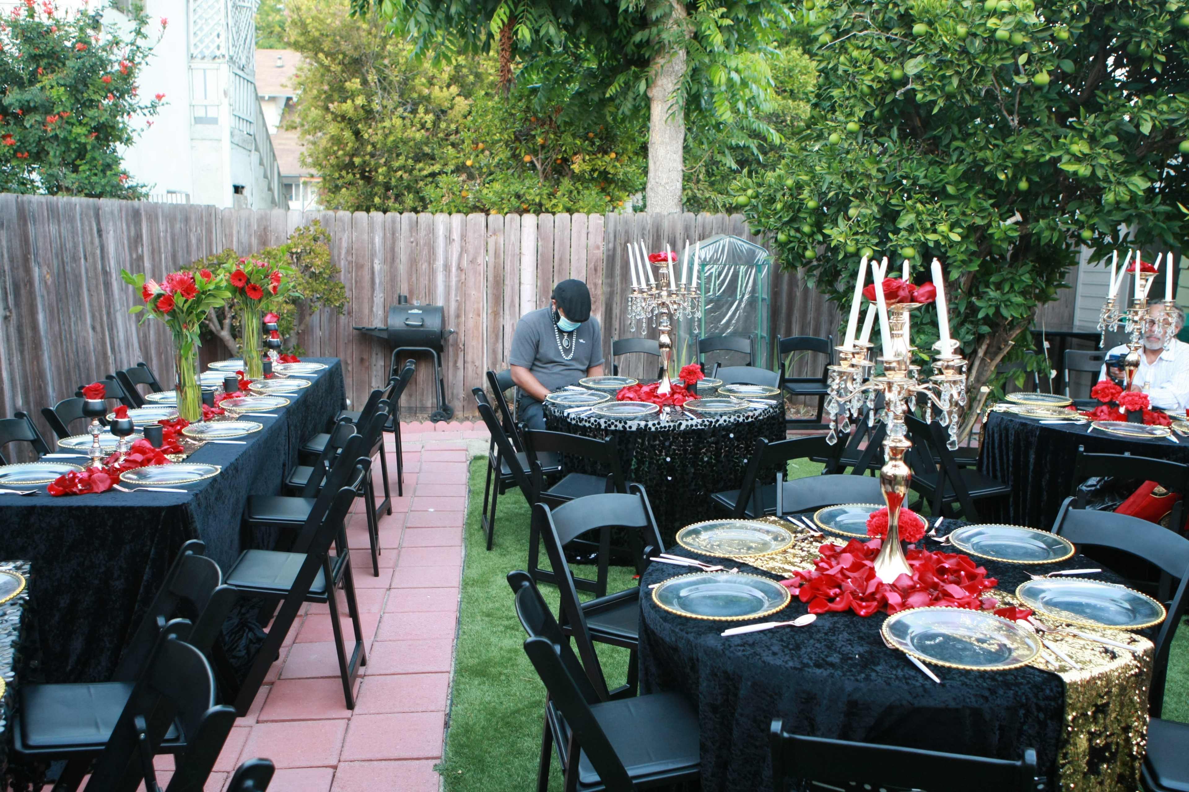 A garden party is set up with several tables adorned in black and red decorations, featuring plates and centerpieces, while a guest sits at one of the tables.
