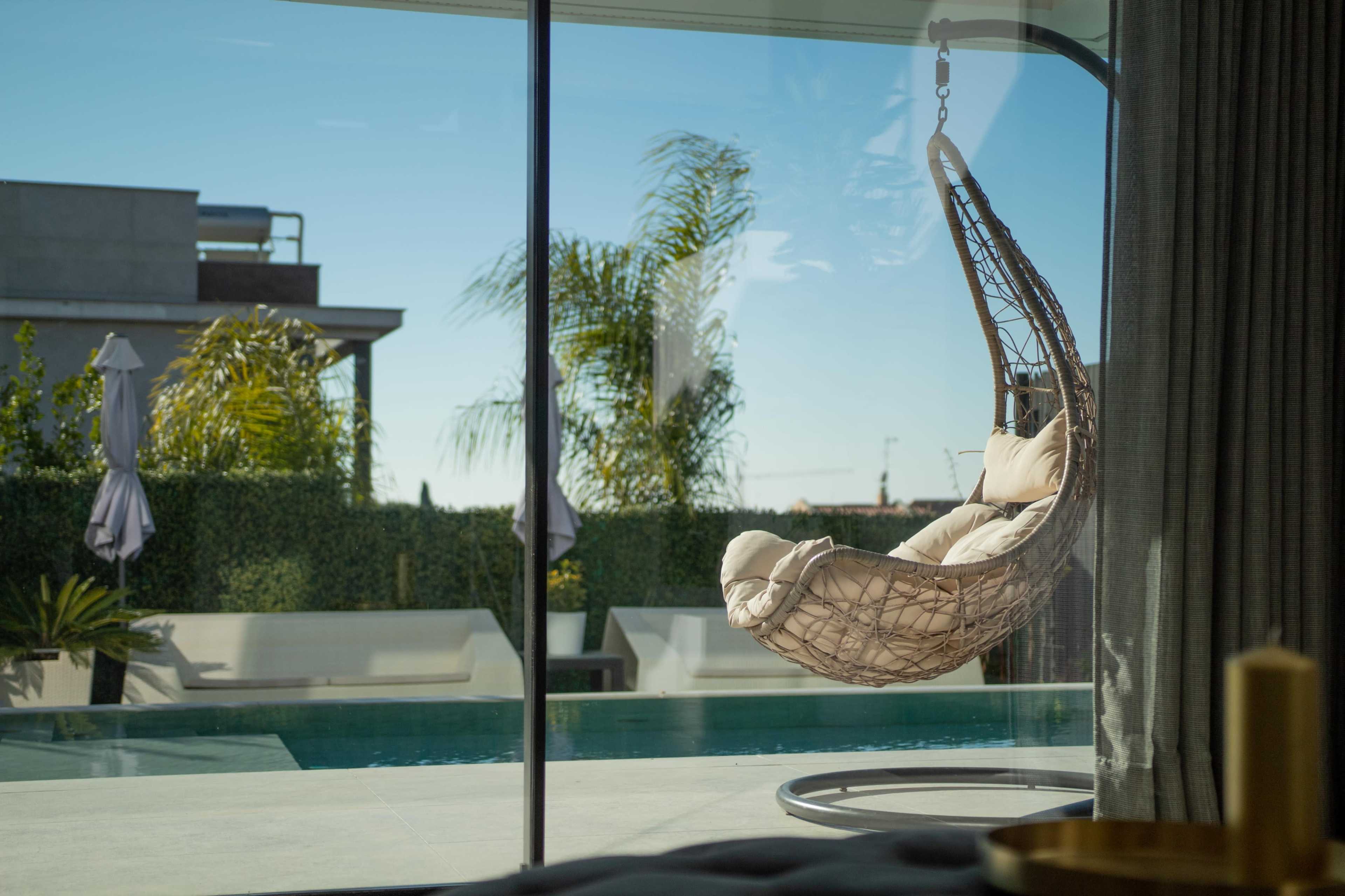 A hanging chair is positioned beside a large glass window overlooking a swimming pool and lush greenery.