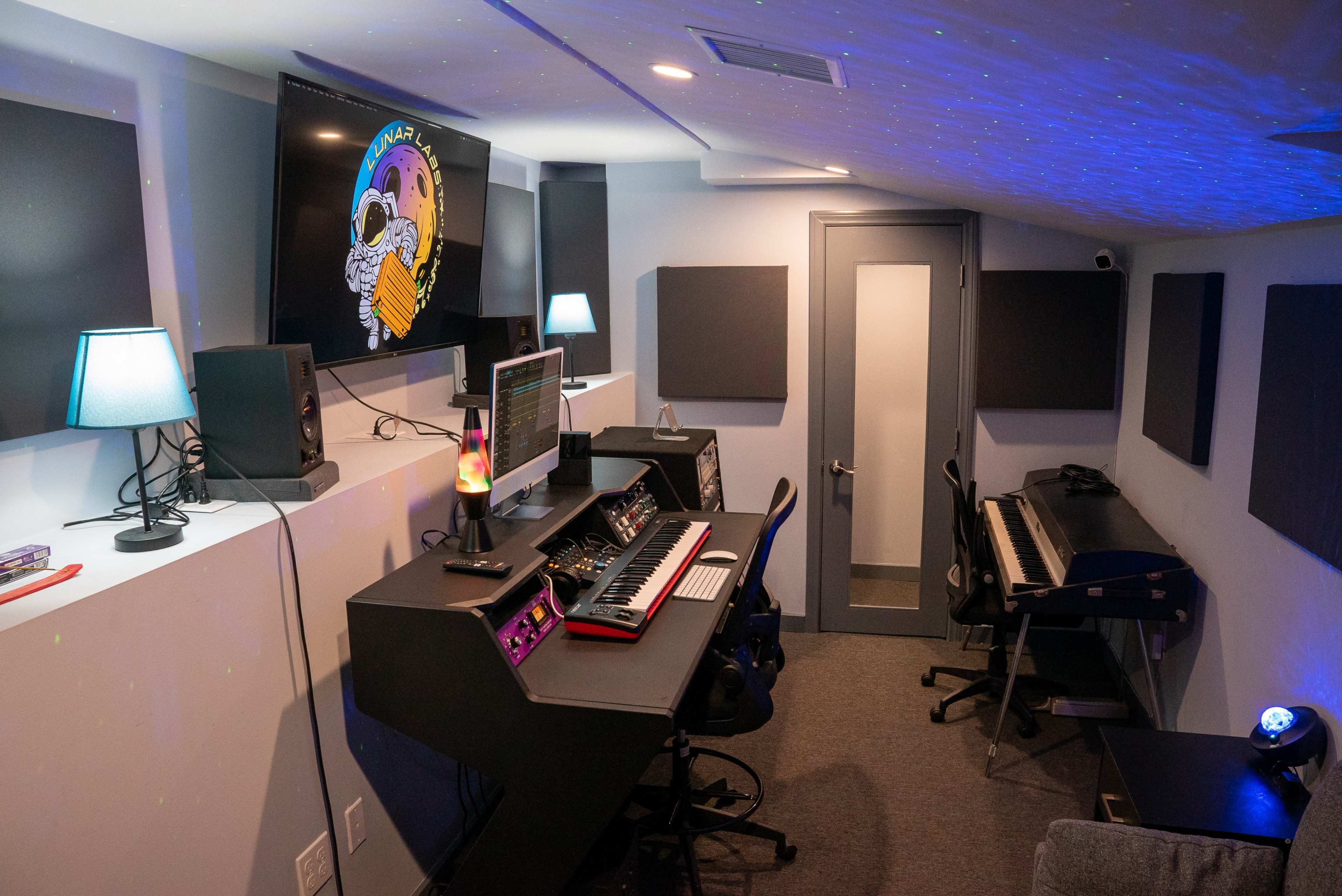 The image shows a music studio equipped with a computer, synthesizers, speakers, and a keyboard, featuring a glowing lamp and soundproof panels on the walls.