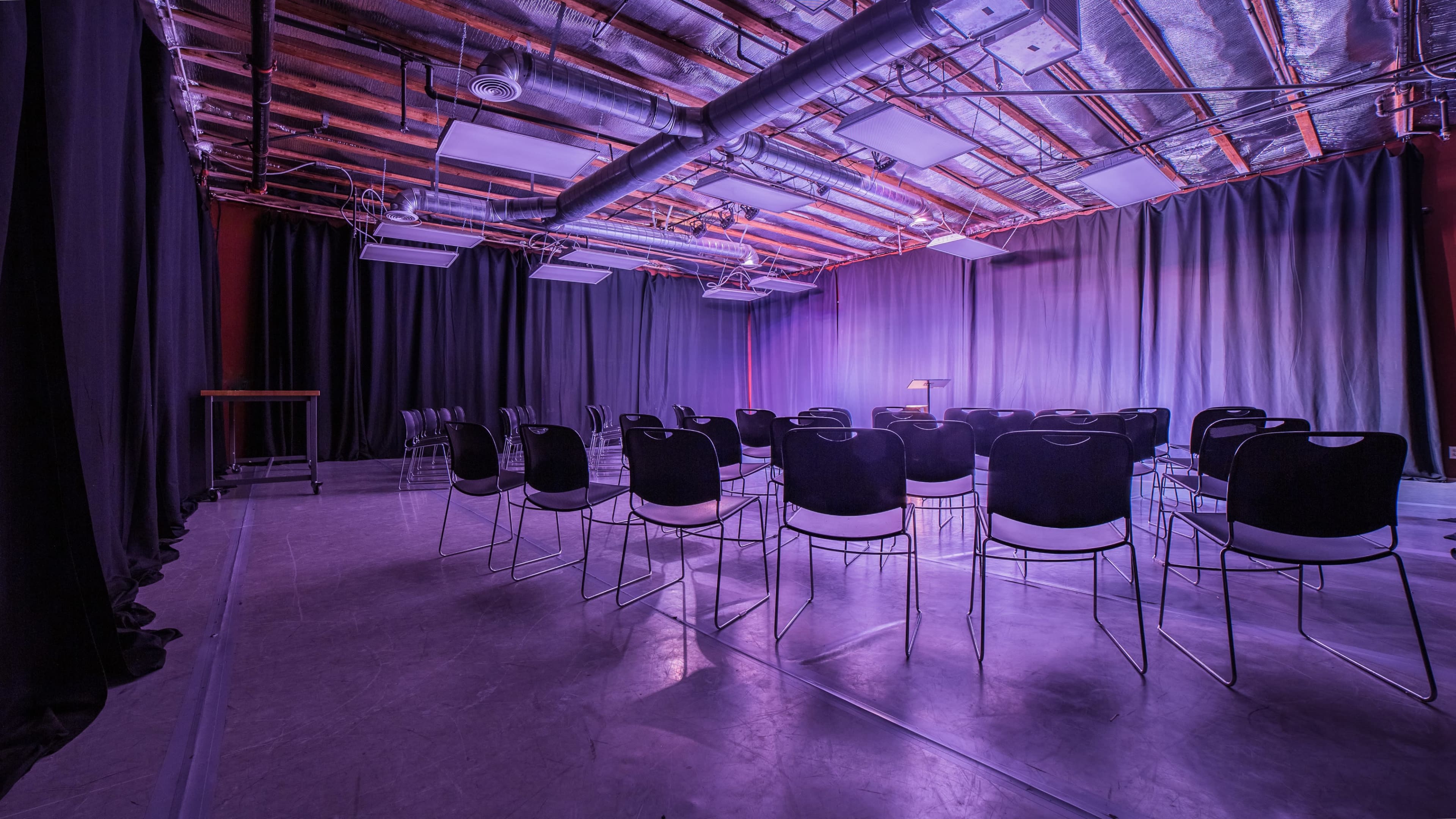 An empty room with rows of black chairs facing a small podium, illuminated by purple lighting.