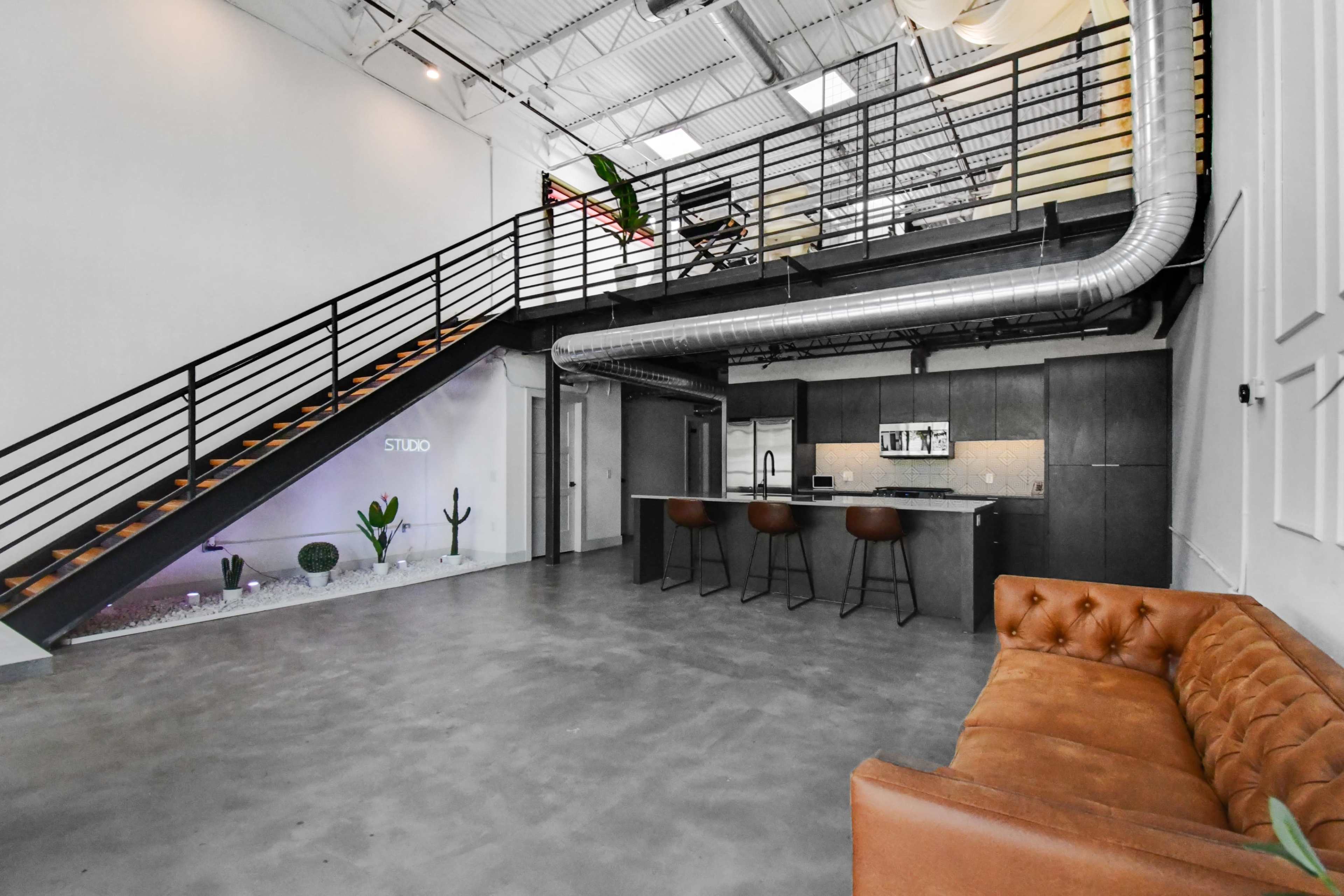 The image shows a modern, industrial-style interior featuring a staircase leading to a loft space, a kitchen area with bar seating, and a couch in the foreground.
