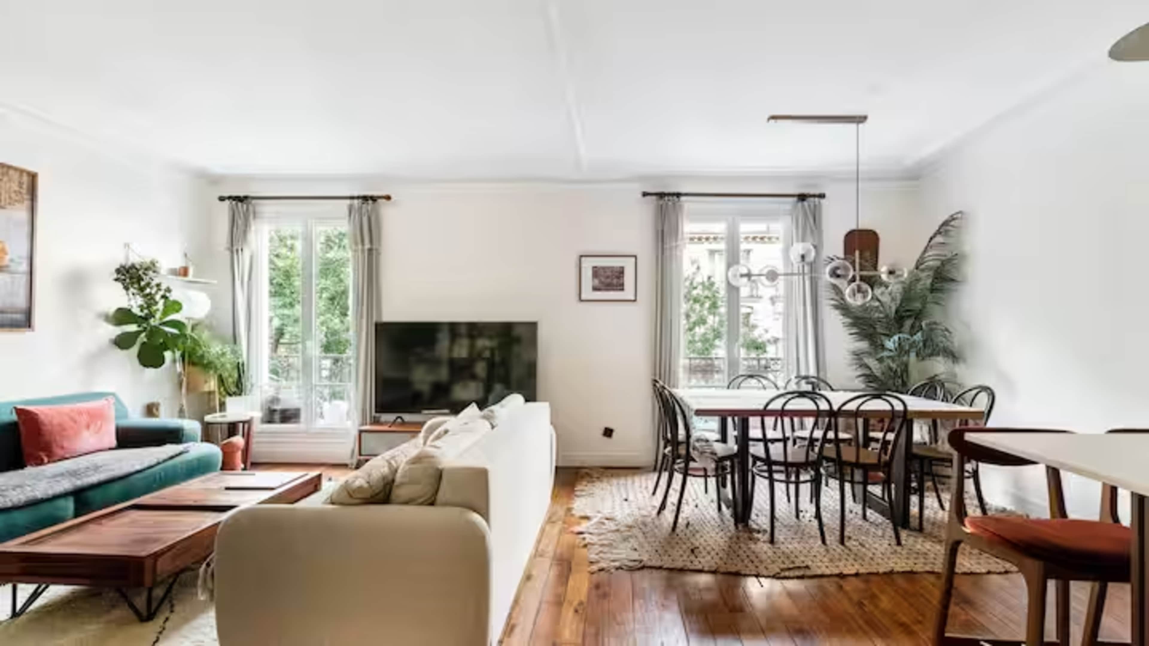 The image shows a bright living room with a sofa, a coffee table, a dining area, and large windows overlooking greenery.