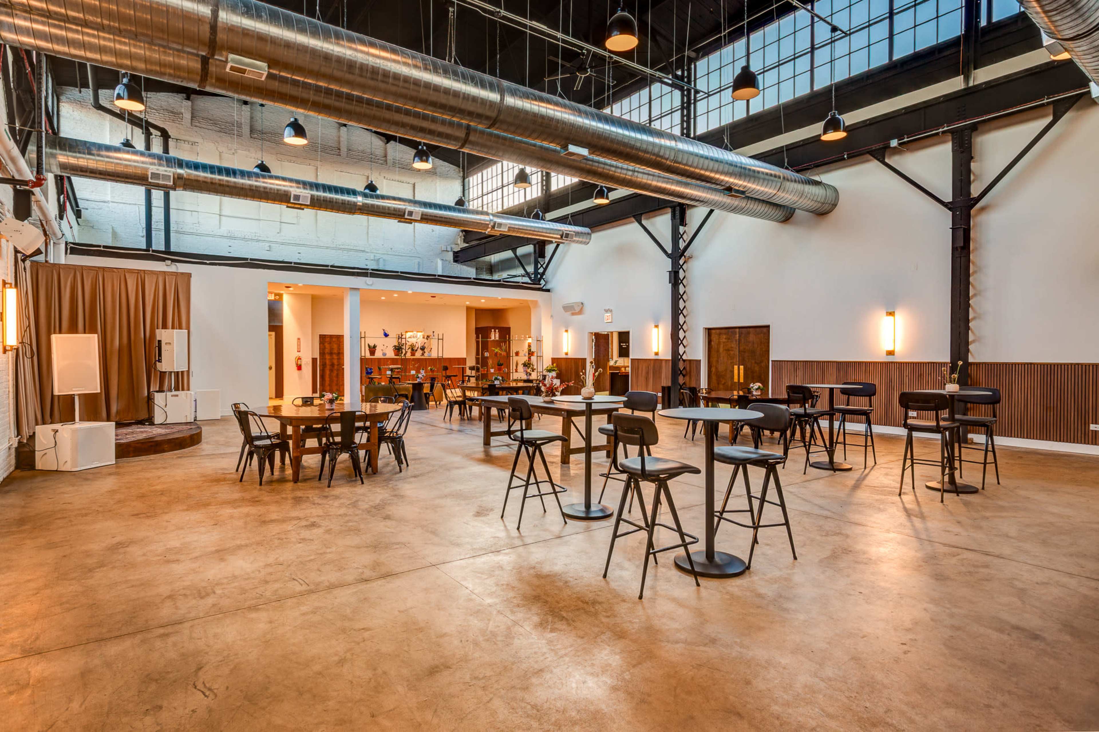 The image shows a spacious interior of a venue featuring high ceilings, large windows, and a mix of tables and chairs arranged throughout the area.