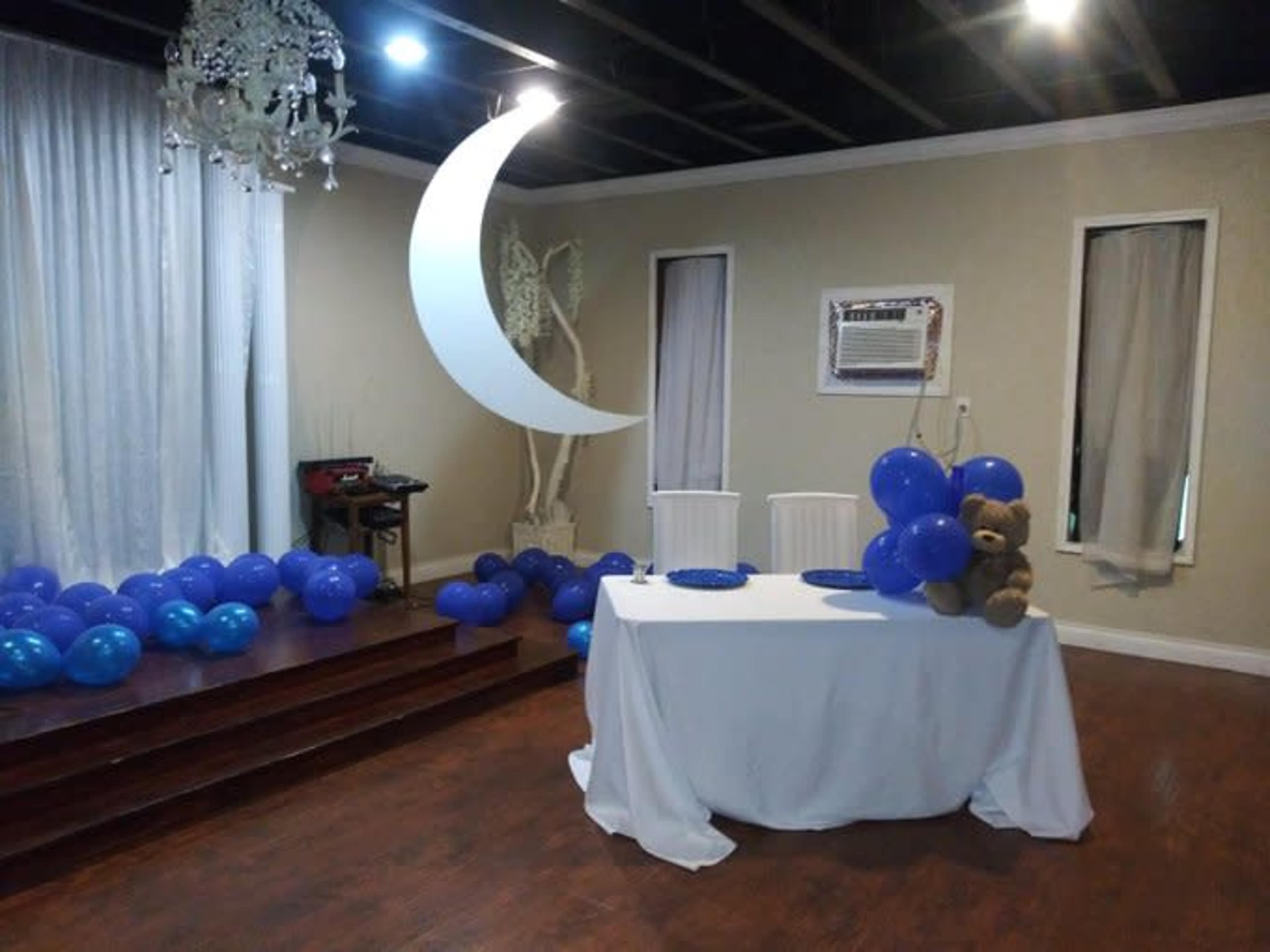 A decorated event space features a white table set with blue plates, surrounded by blue balloons and a teddy bear, beneath a decorative crescent moon and curtain backdrop.