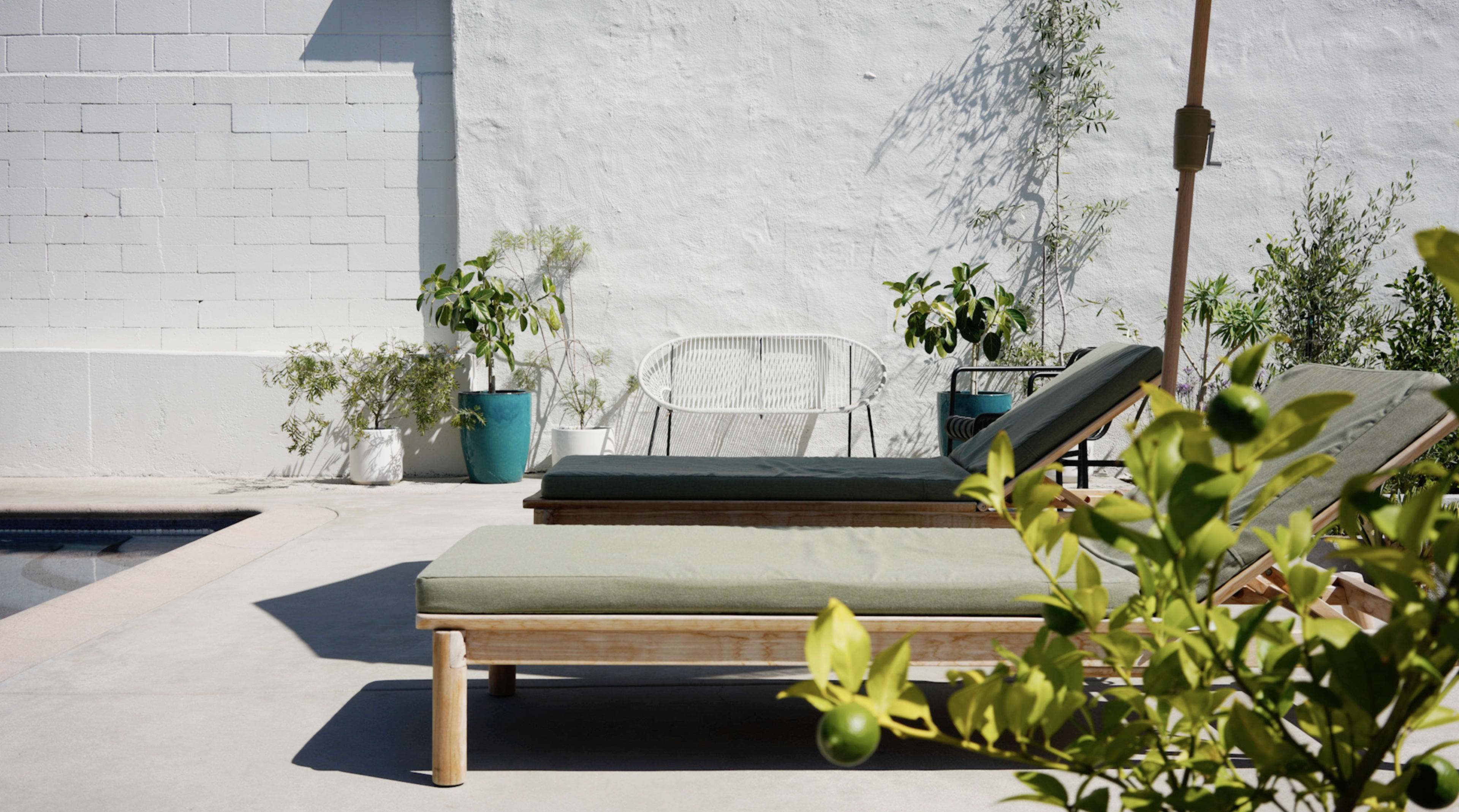 Two green lounge chairs are positioned beside a pool area, with potted plants and a white chair against a light-colored wall.