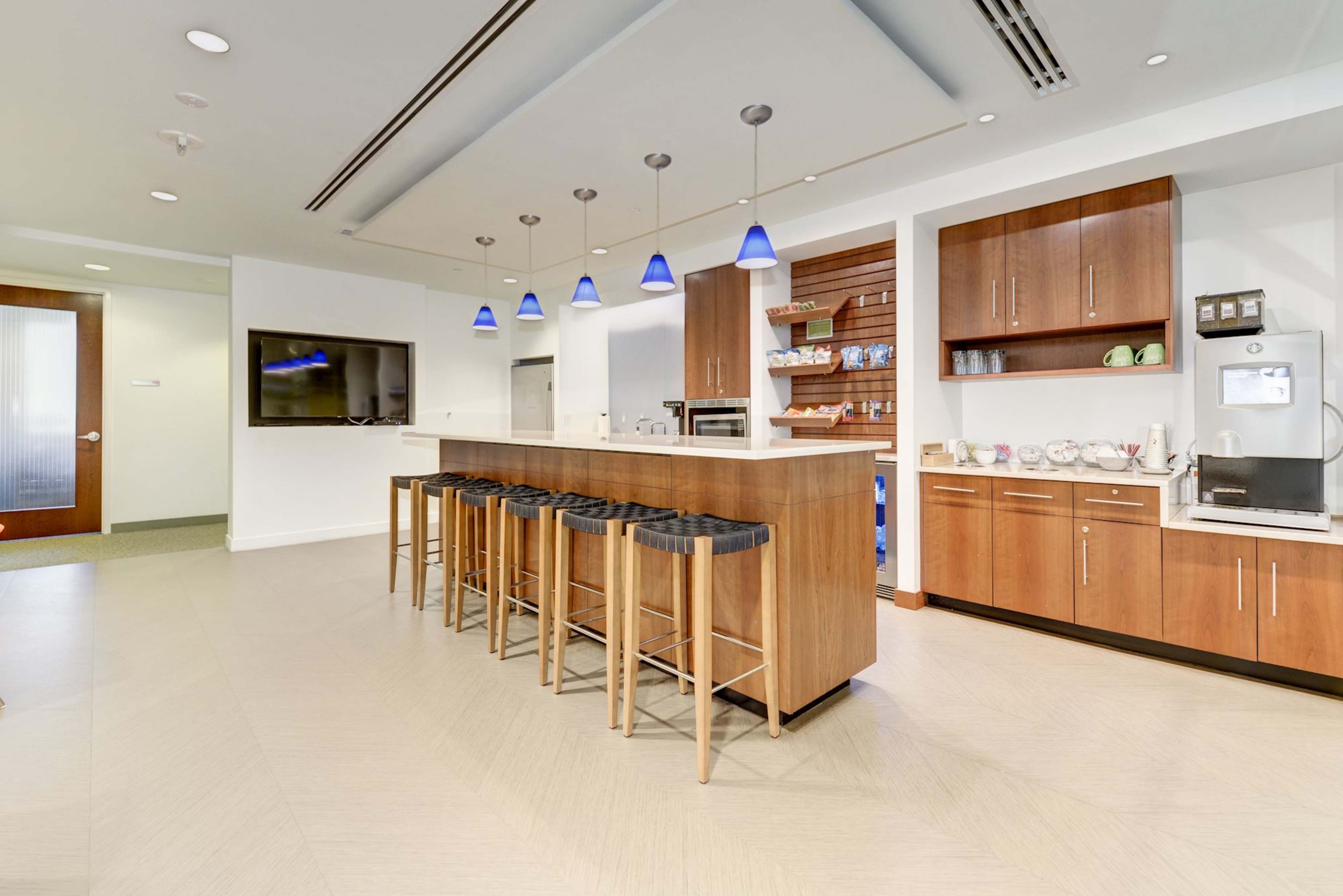A modern kitchen area features a long island with bar stools, overhead lights, and a coffee station against a wall of cabinets.
