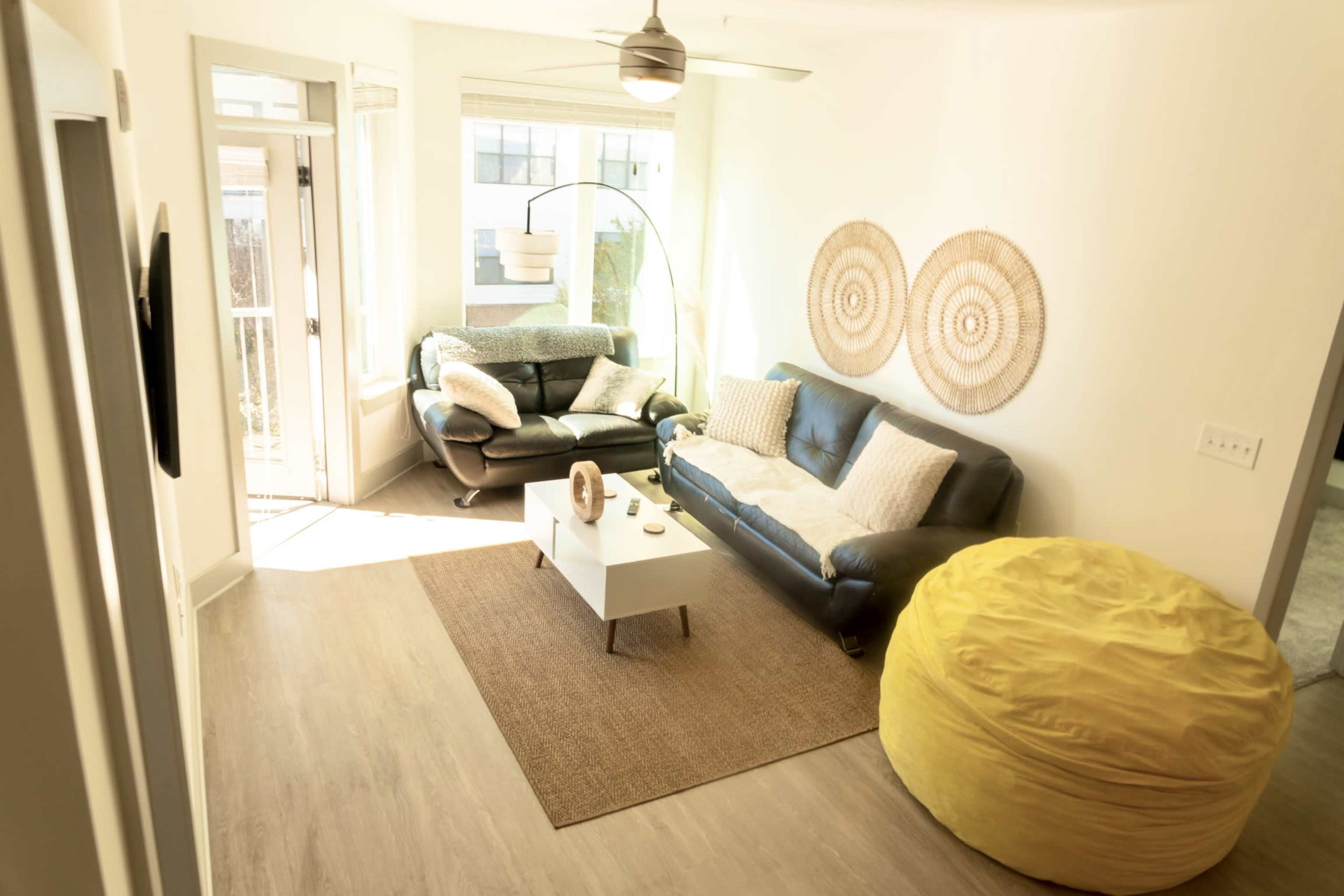 A bright living room with a leather sofa, a round coffee table, a large yellow bean bag, and decorative wall art made of woven materials.