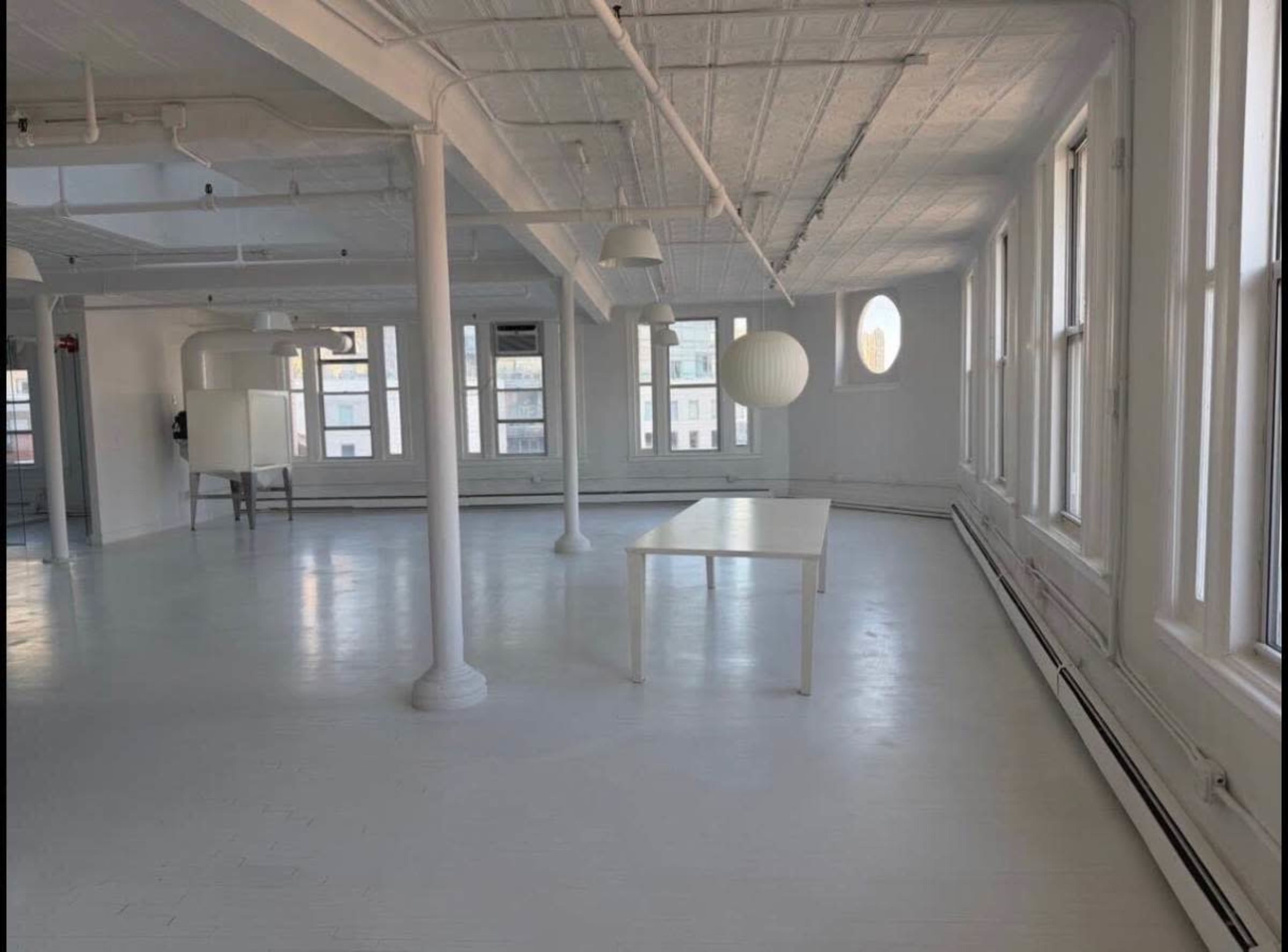 The image shows a spacious, well-lit room with white walls and a large table in the center, surrounded by several windows.