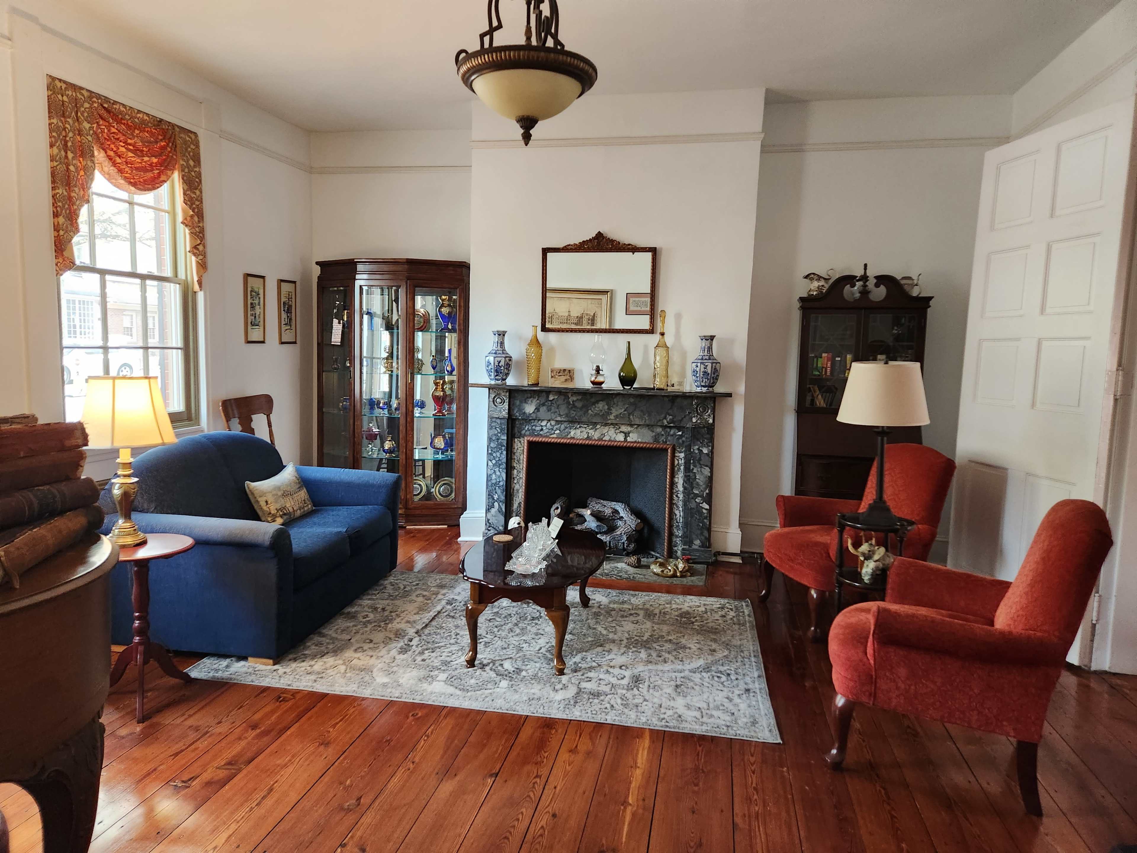 A living room with a blue couch, two red armchairs, a wooden coffee table on a patterned rug, and a fireplace with decorative items.
