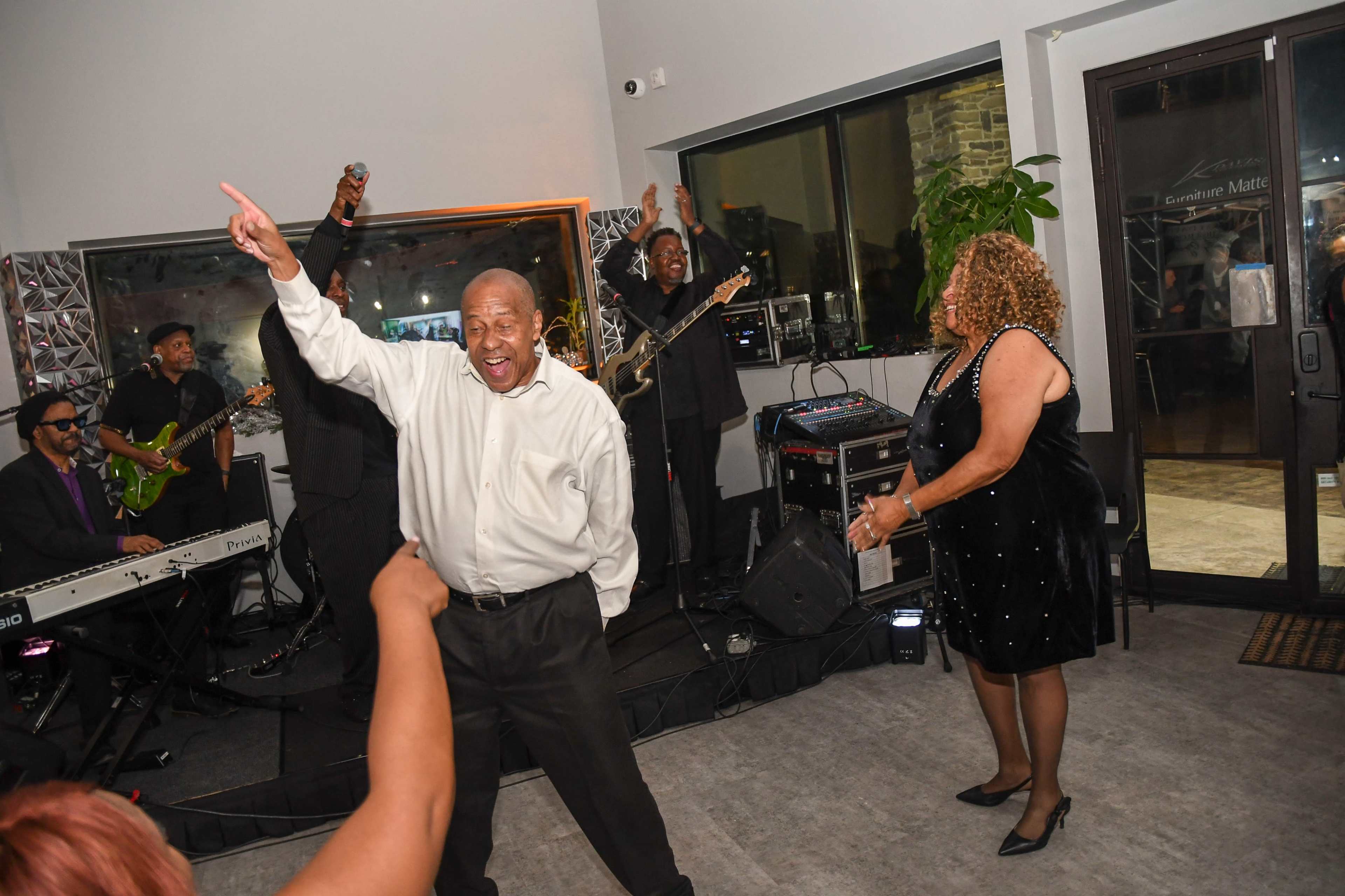 A man in a white shirt dances joyfully in front of a live band while a woman in a black dress watches and smiles.