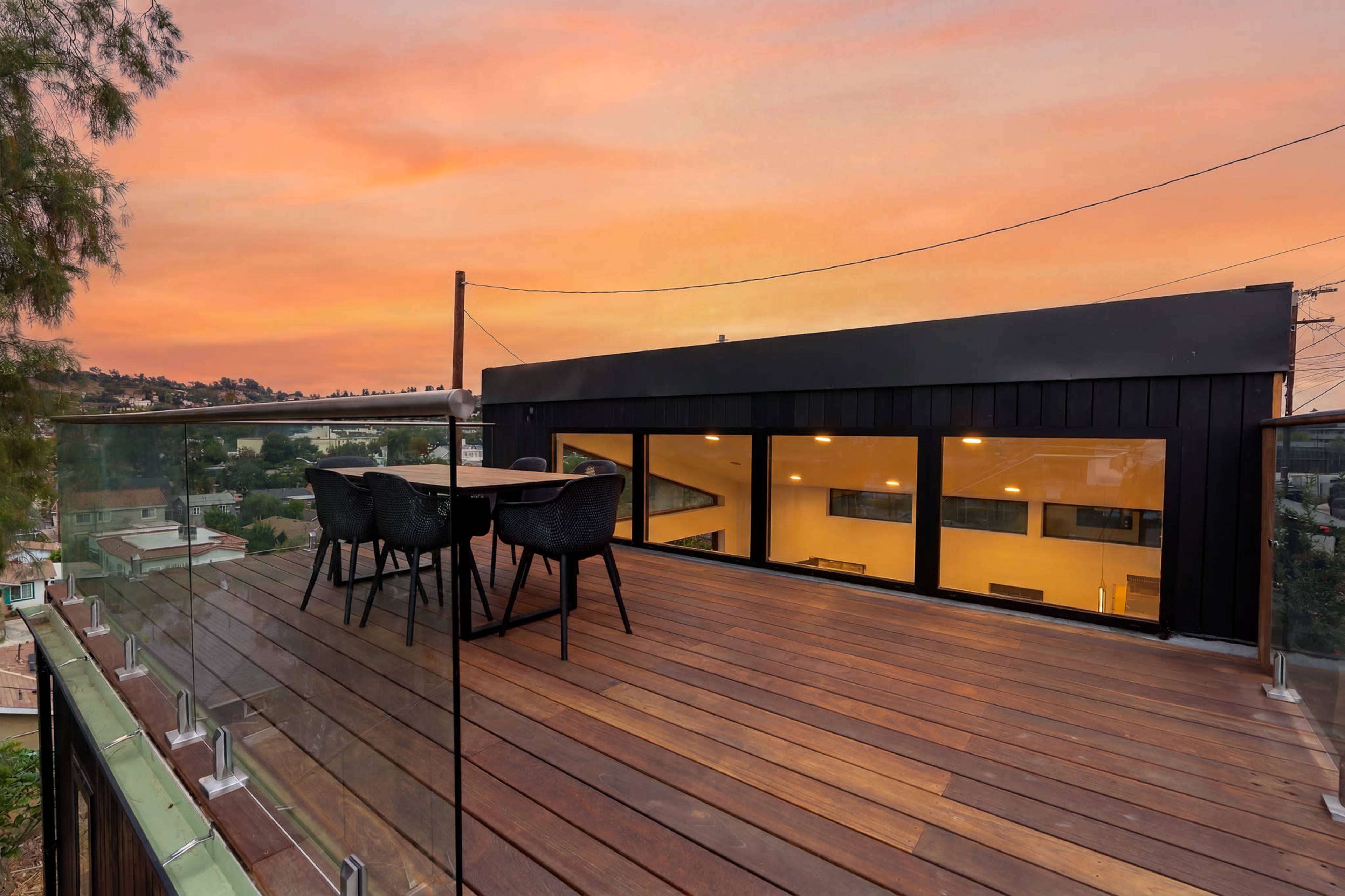 A rooftop deck with a wooden floor features a table and chairs, overlooking a scenic area at sunset.