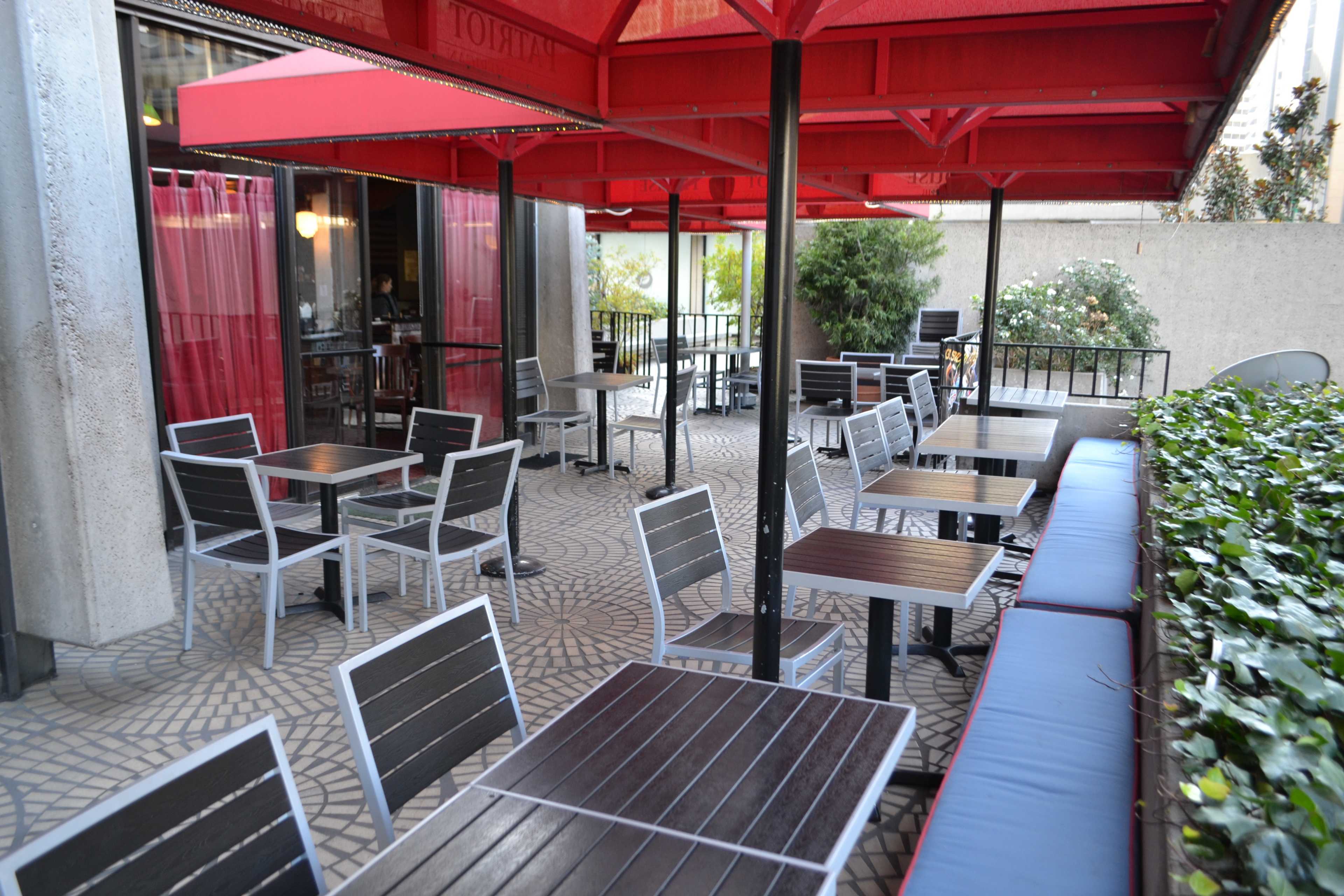 An outdoor dining area with wooden tables and chairs is shaded by red umbrellas.