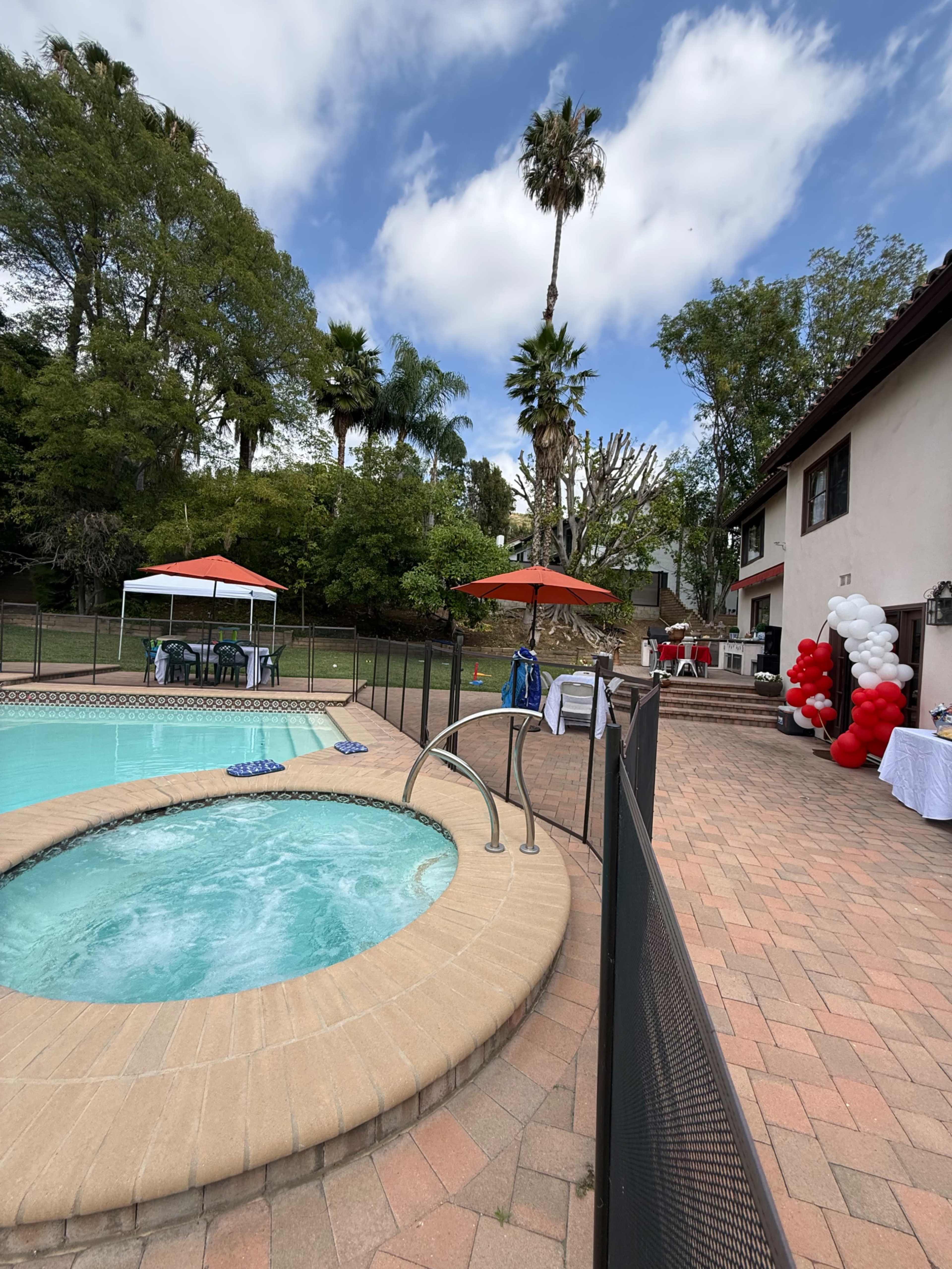A pool area features a hot tub, umbrellas, and a decorative setup with balloons near a house surrounded by trees.
