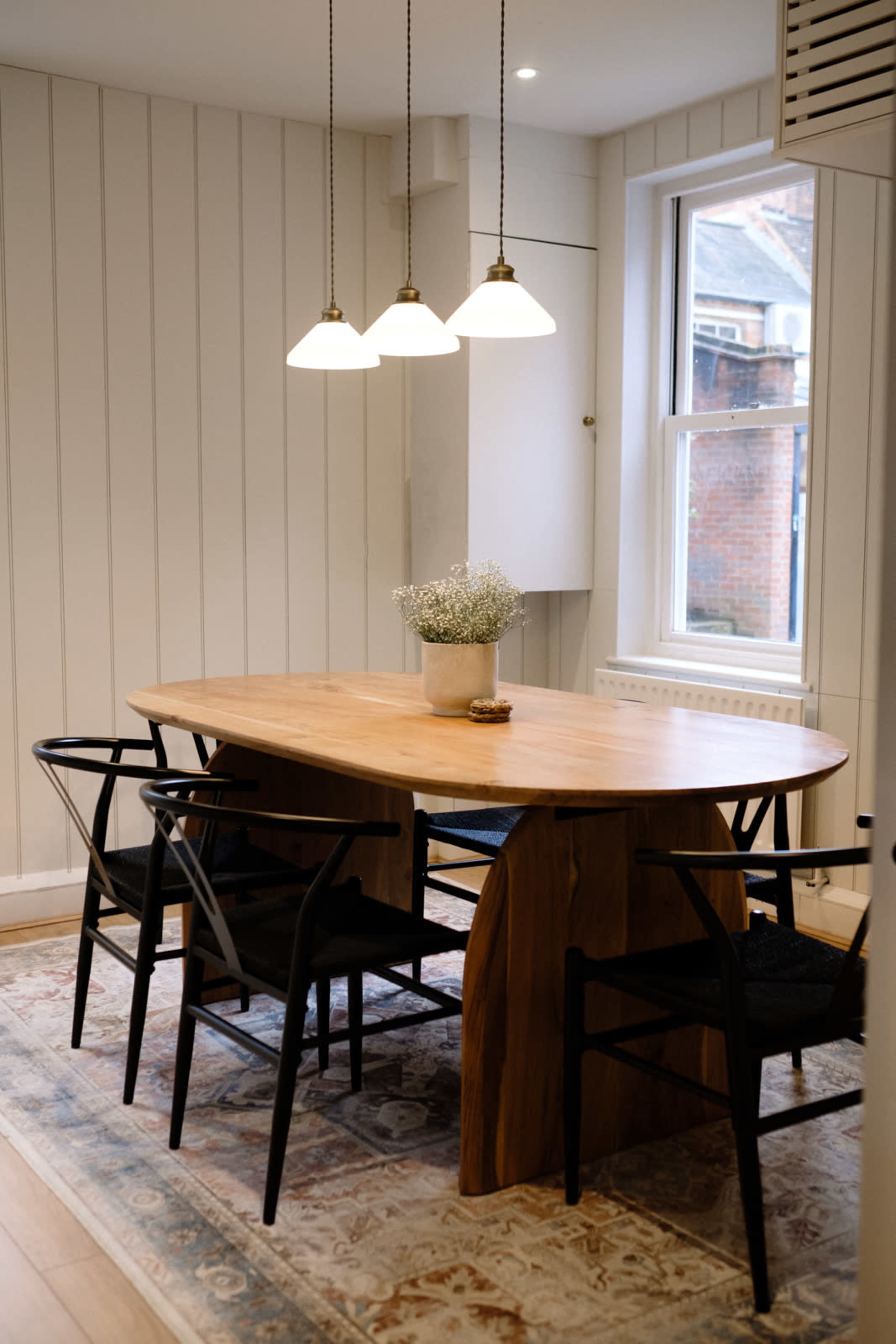 A wooden oval dining table surrounded by black chairs sits under three pendant lights in a well-lit room with white paneling.