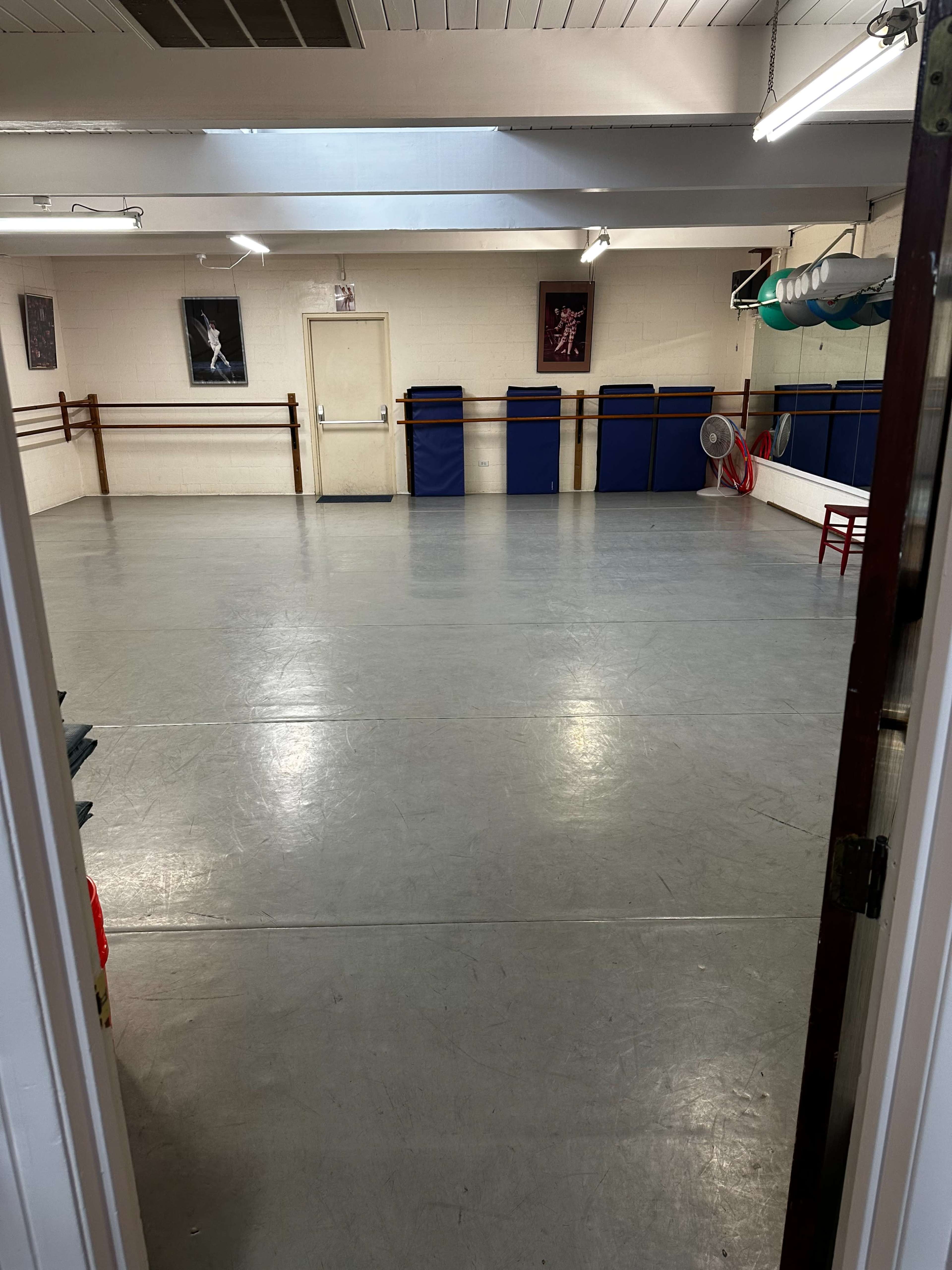 The image shows an empty dance studio with a large, open floor, mirrored walls, and some equipment neatly arranged along the sides.