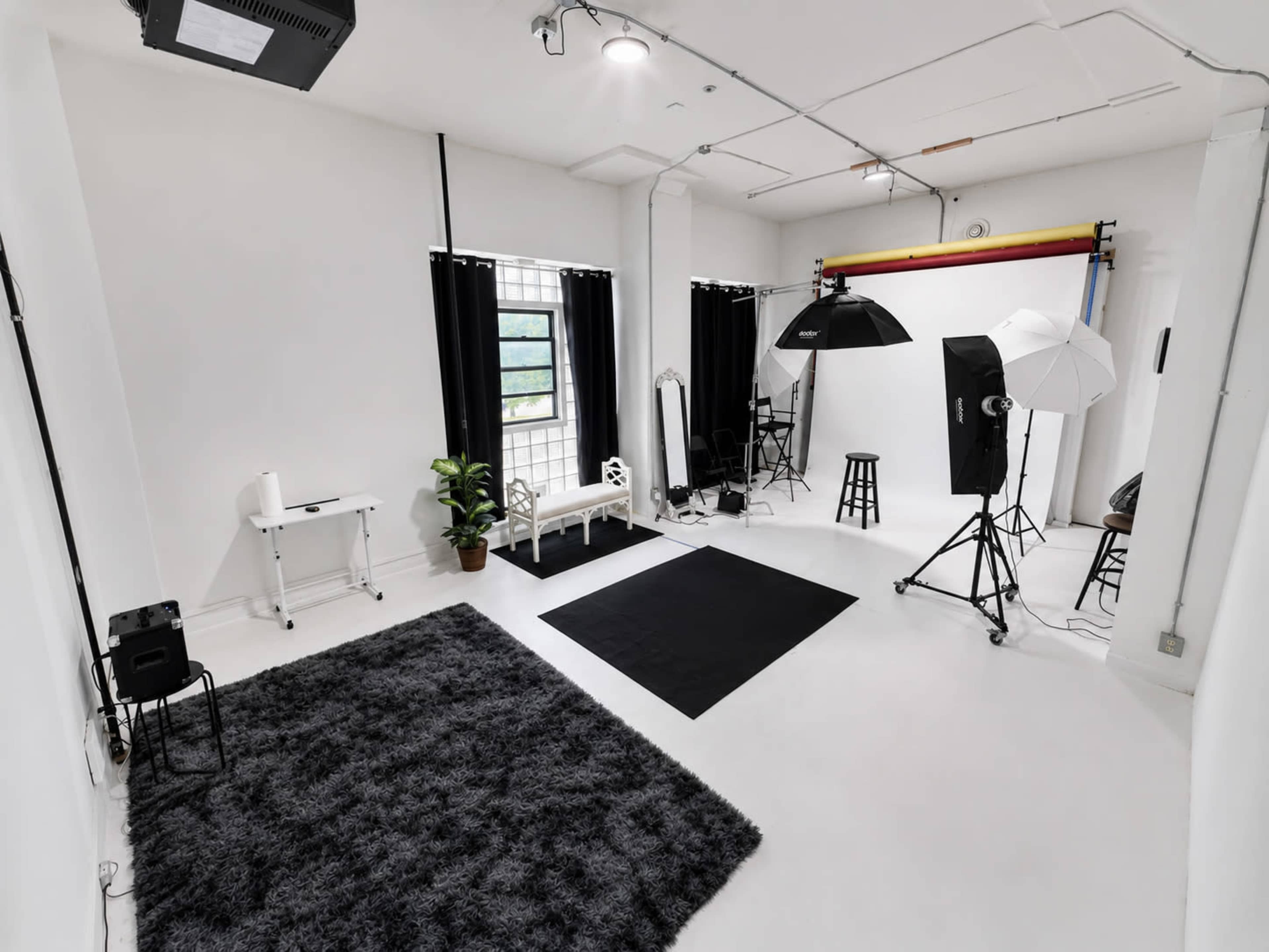The image shows a spacious photography studio featuring white walls, two large softbox lights, a black rug, and a variety of props including a chair and a table.