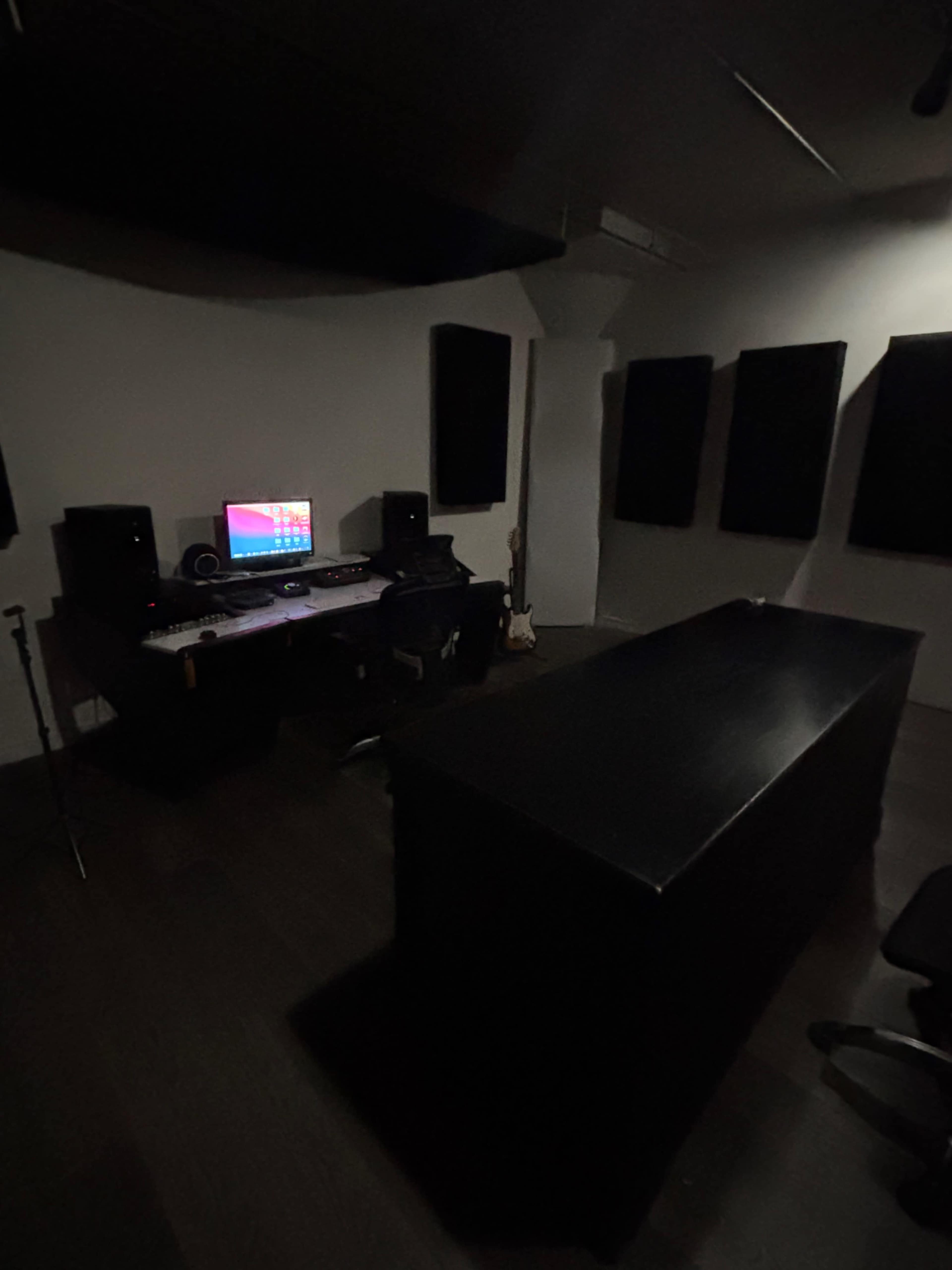 The image shows a dimly lit music studio featuring a desk with a computer, speakers, and soundproof panels on the walls.