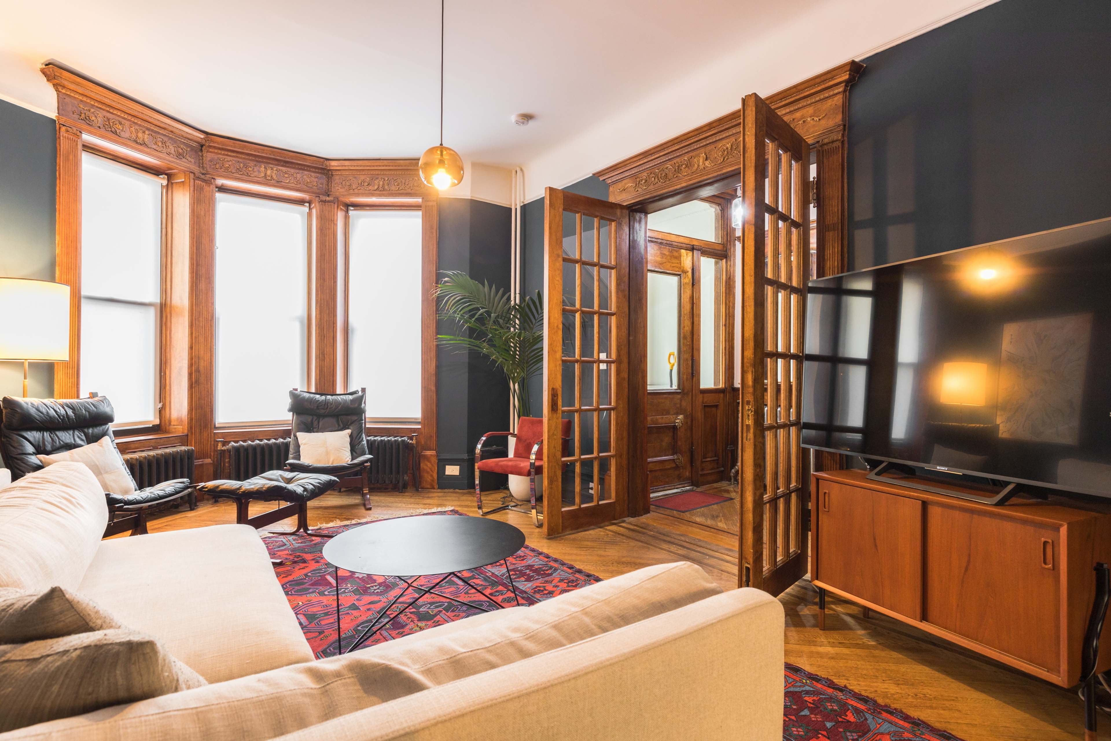 A living room featuring a beige couch, black chairs, a decorative rug, and a large television, with wooden framed doors leading to an adjacent area.