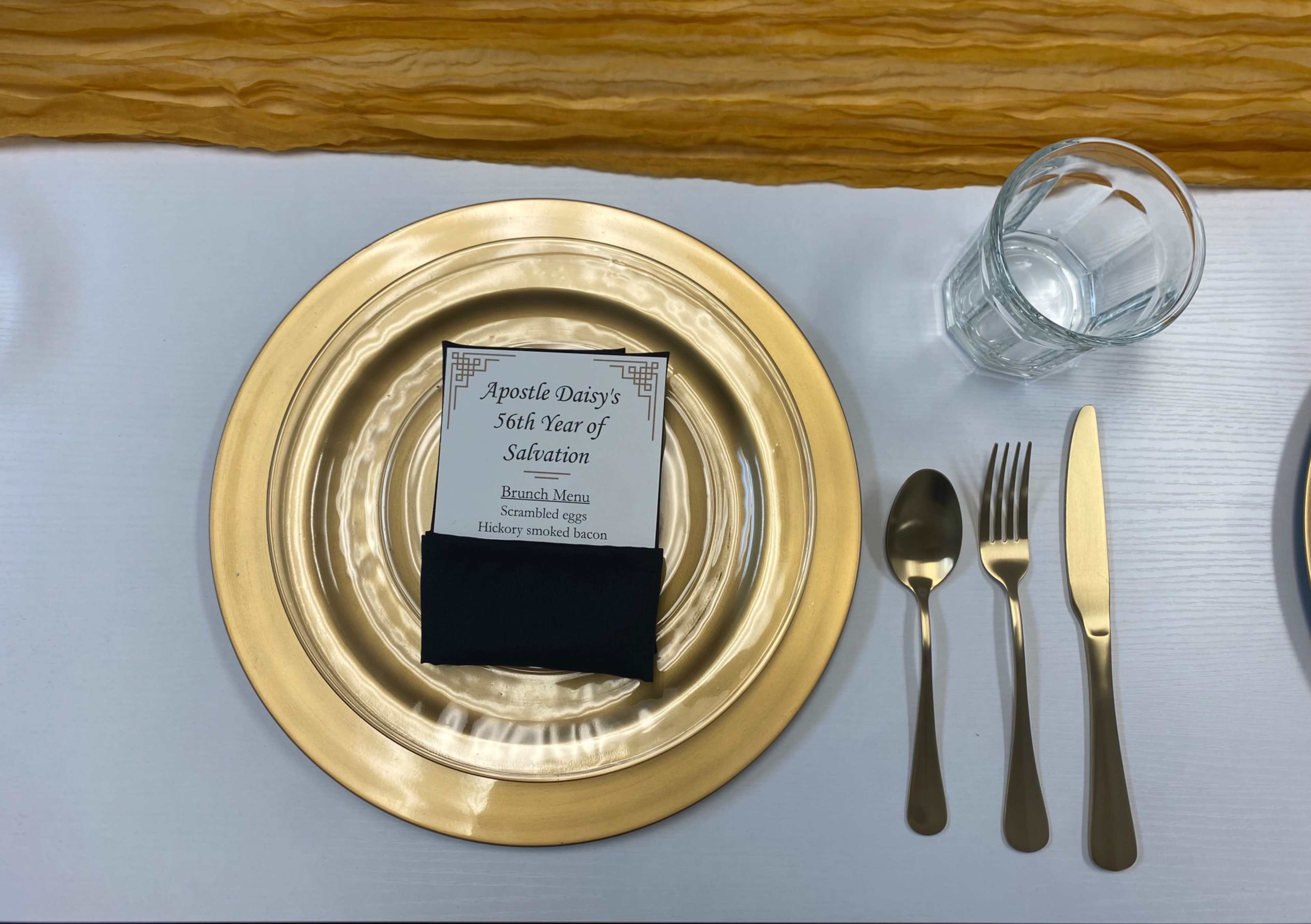 The image shows a dining table setting with a gold charger plate, a menu card placed on a dark napkin, and utensils arranged neatly beside a glass of water.