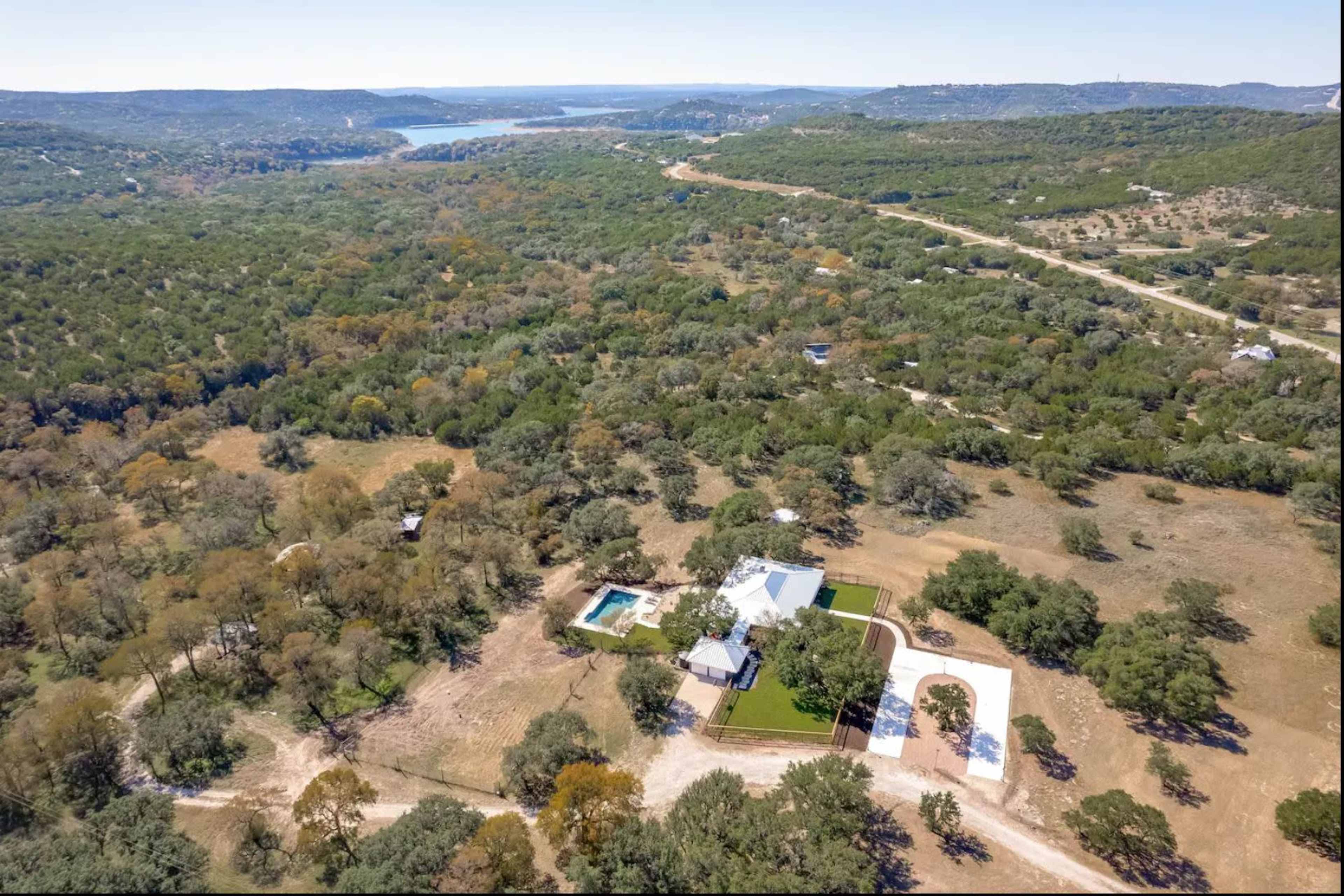 The image shows an aerial view of a spacious property with a house, pool, and landscaped areas surrounded by wooded terrain and a distant body of water.