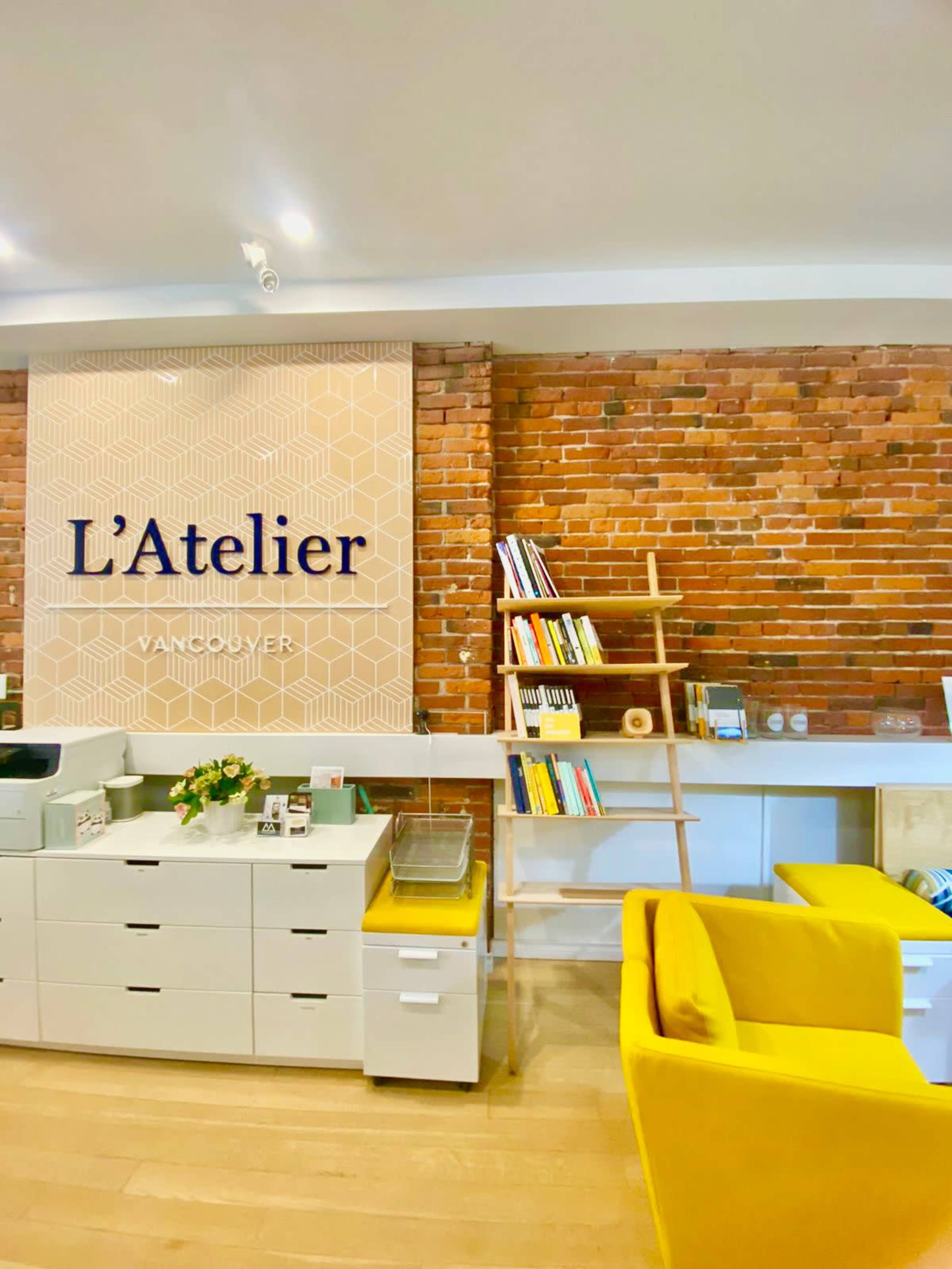 The image shows a modern office interior with a bright yellow chair, a decorative bookshelf, and a brick wall featuring the name "L'Atelier Vancouver."