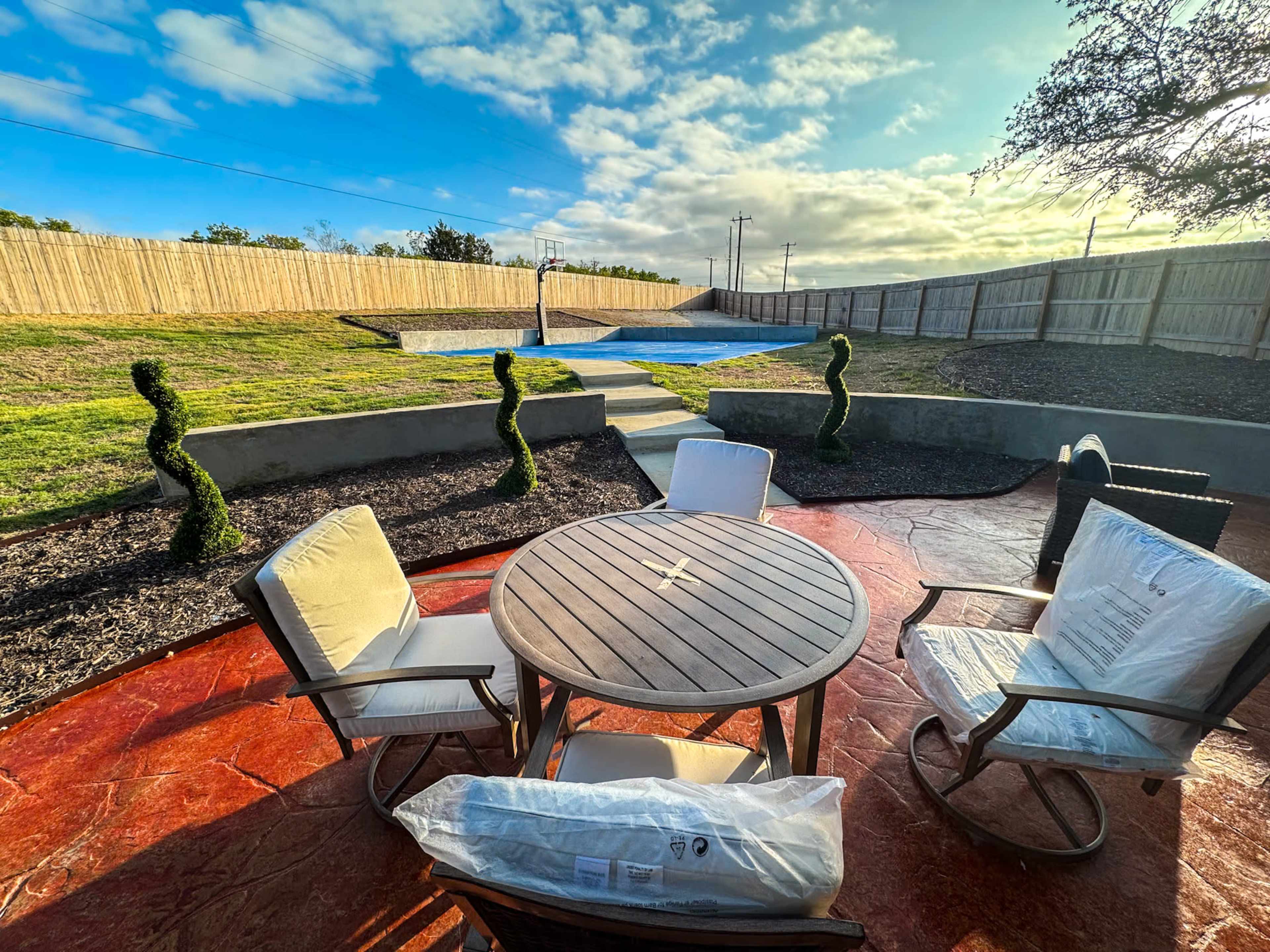 A circular wooden table with four chairs is set on a patio overlooking a lawn and a covered swimming pool, surrounded by neatly trimmed shrubs.