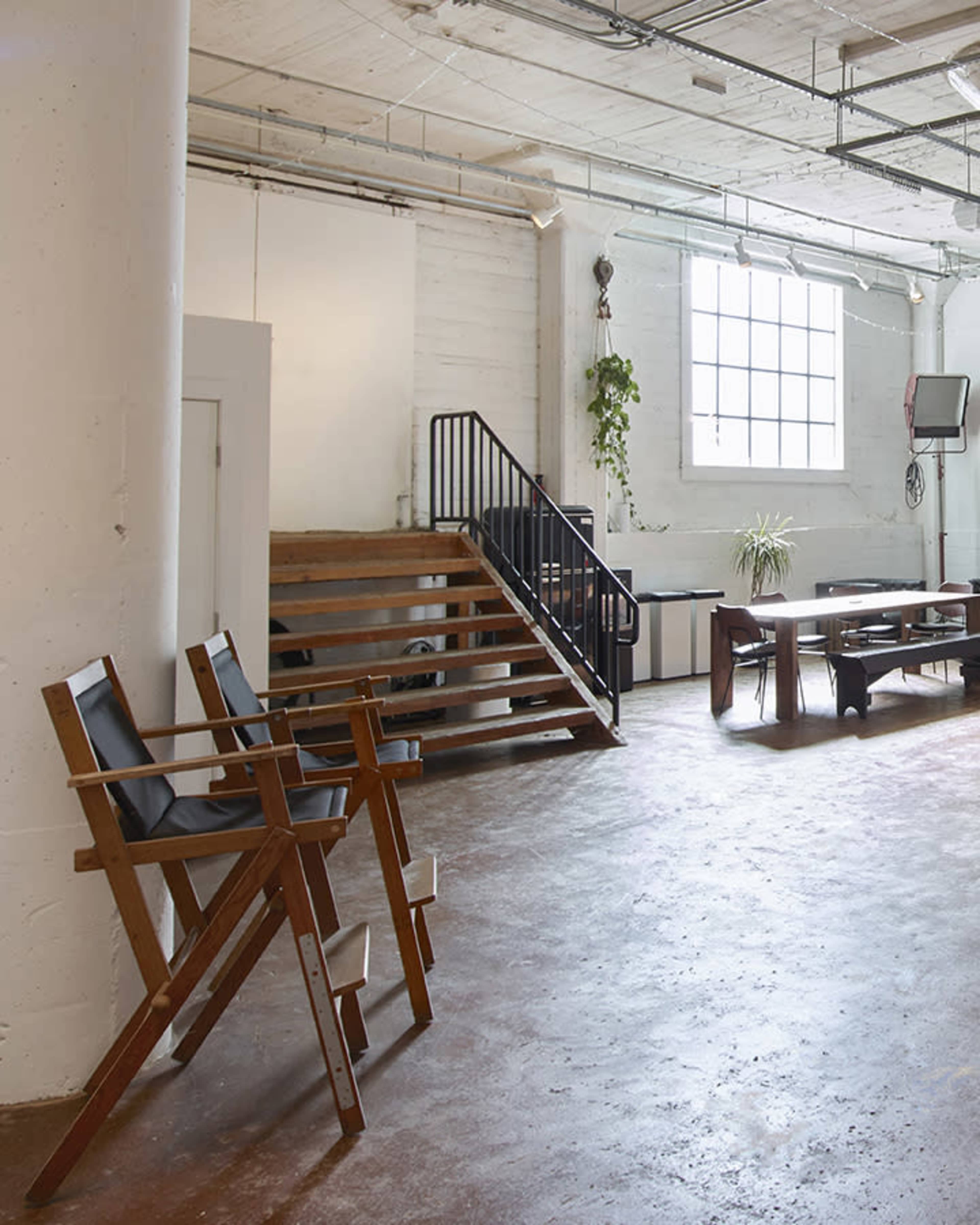 The image shows a well-lit, spacious interior featuring wooden director's chairs, a staircase, and a table with chairs.