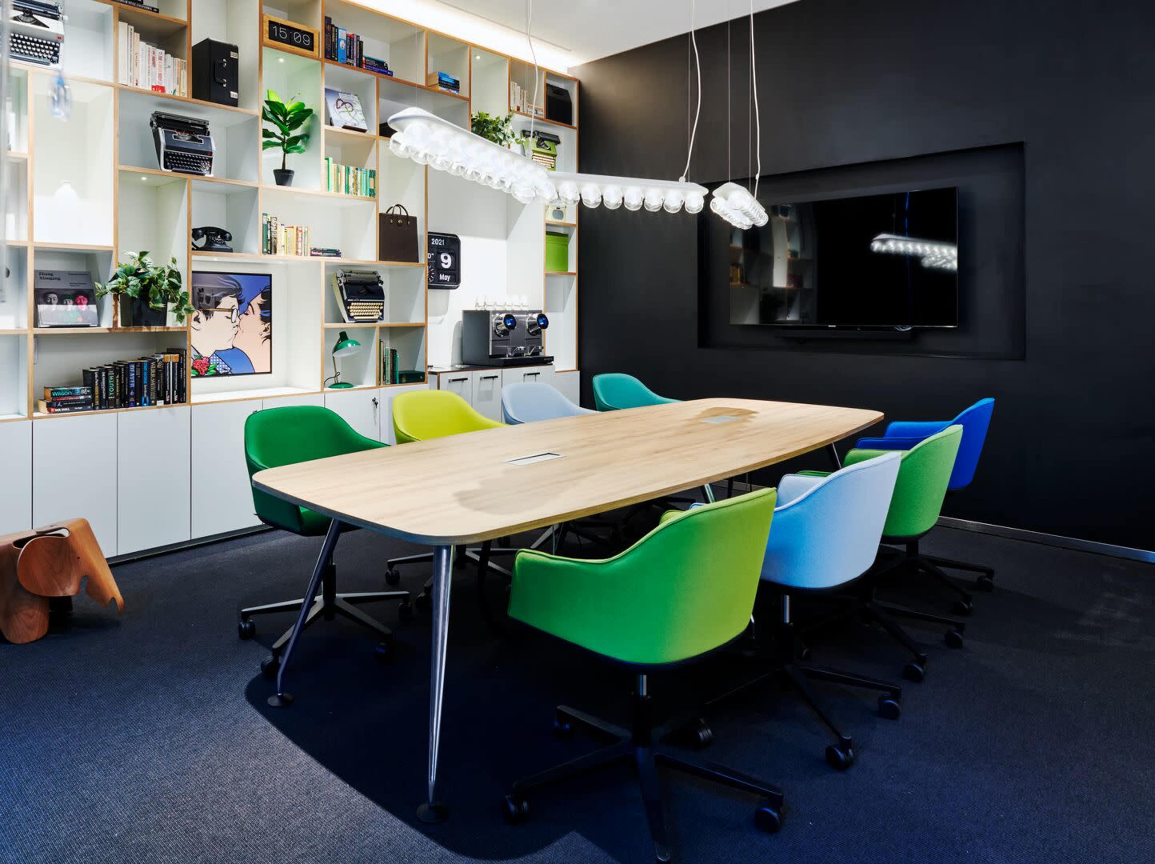 A modern conference room with a long wooden table surrounded by colorful ergonomic chairs and a large wall-mounted screen.