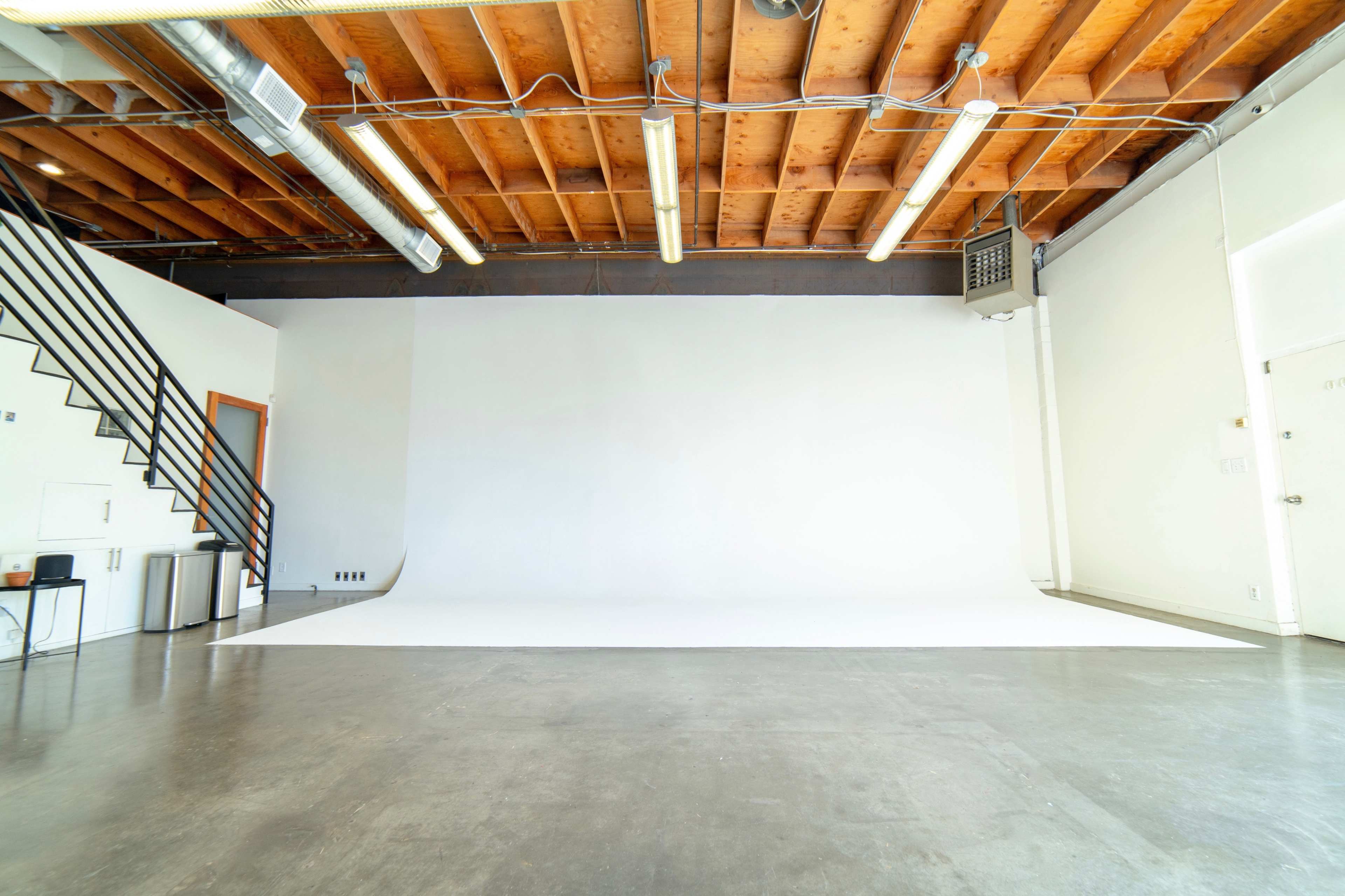 A spacious, empty studio with a white backdrop and wooden ceiling beams, featuring a staircase and polished concrete floor.
