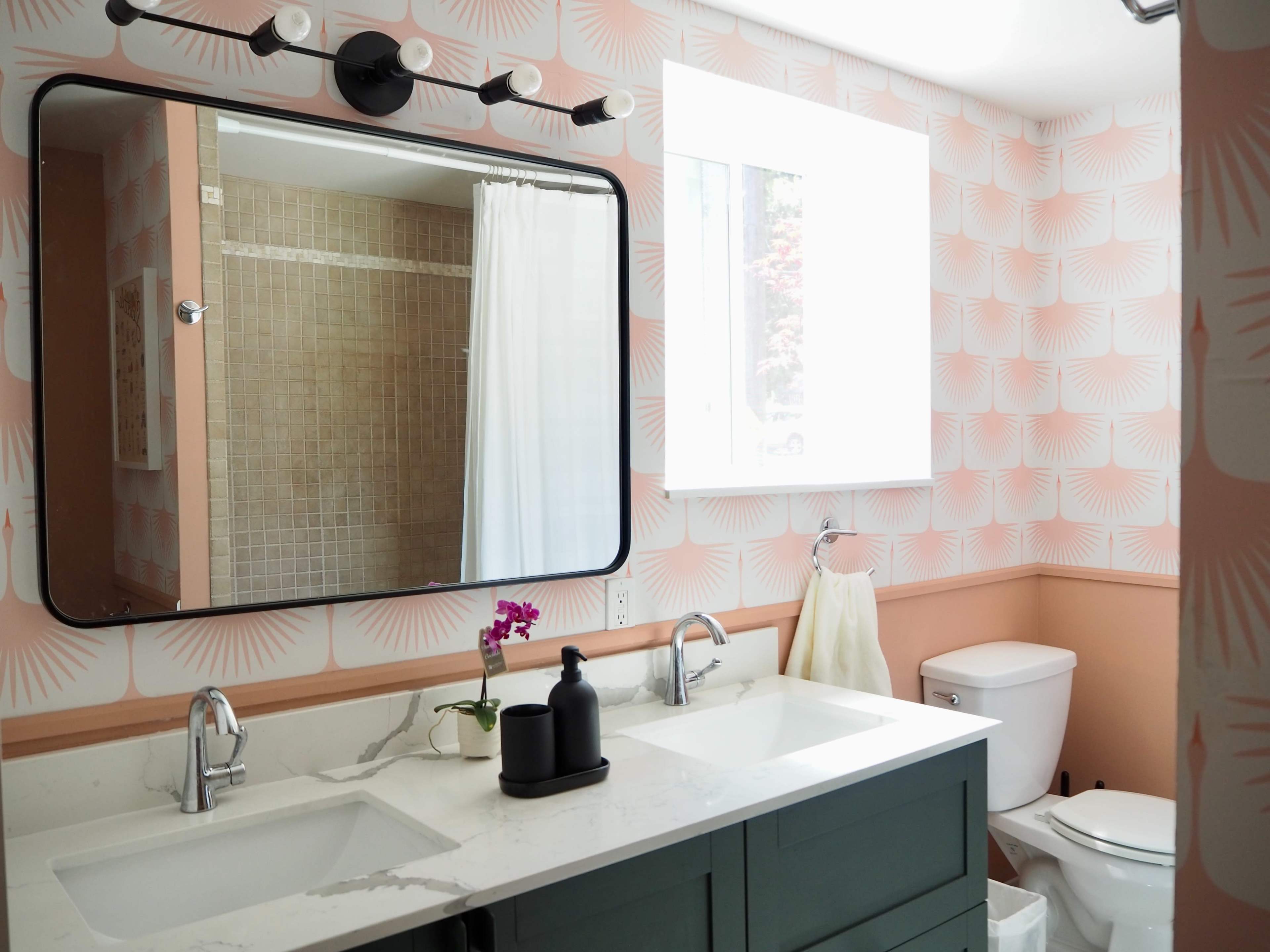 A modern bathroom features dual sinks with a large mirror, a shower area with beige tiles, and light peach-colored wallpaper adorned with simple patterns.